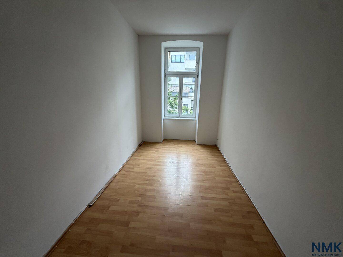 Schmaler Raum mit hellem Laminatboden und einem hohen Fenster, ideal als Schlafzimmer.