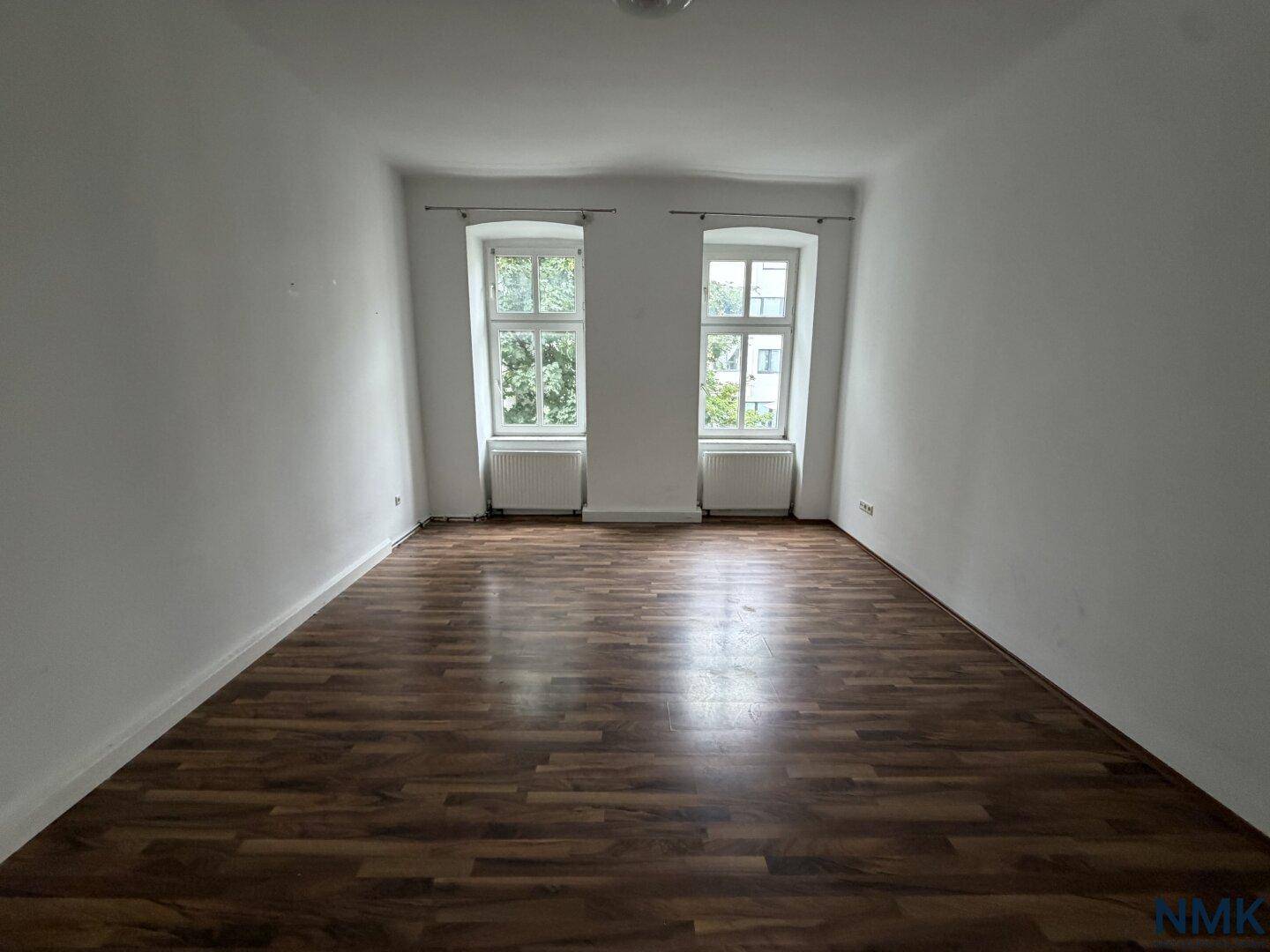 Leeres Zimmer mit dunklem Laminatboden und zwei Fenstern, bereit zur Gestaltung.