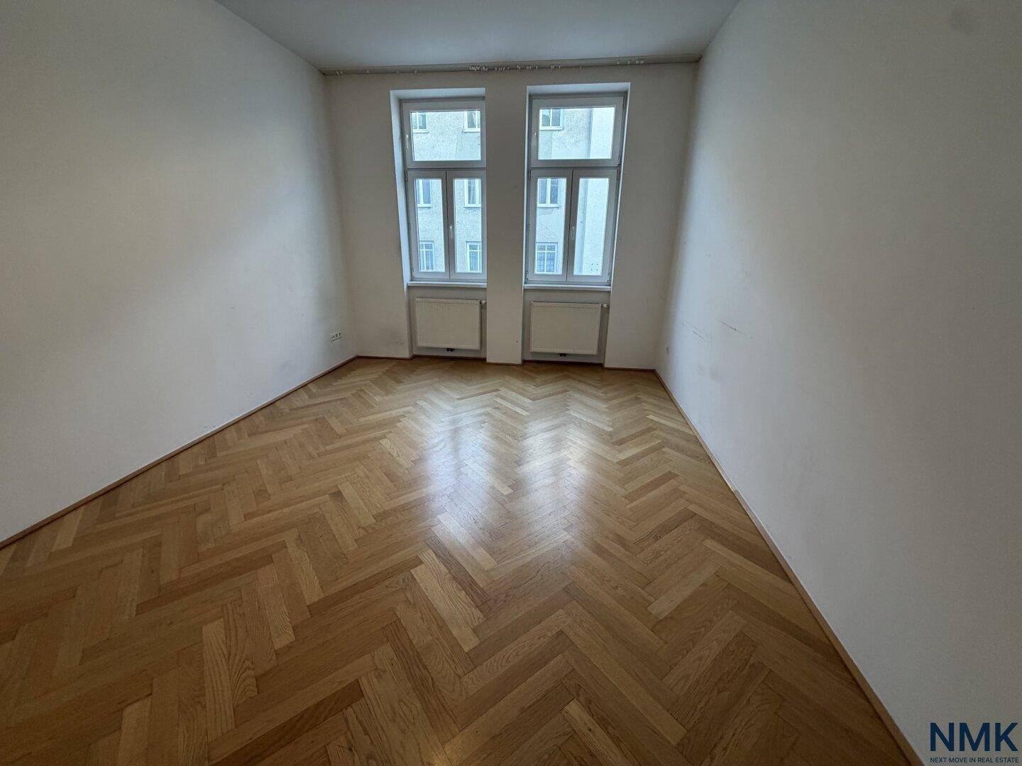Leeres Zimmer mit klassischem Fischgrätparkett und zwei großen Fenstern.