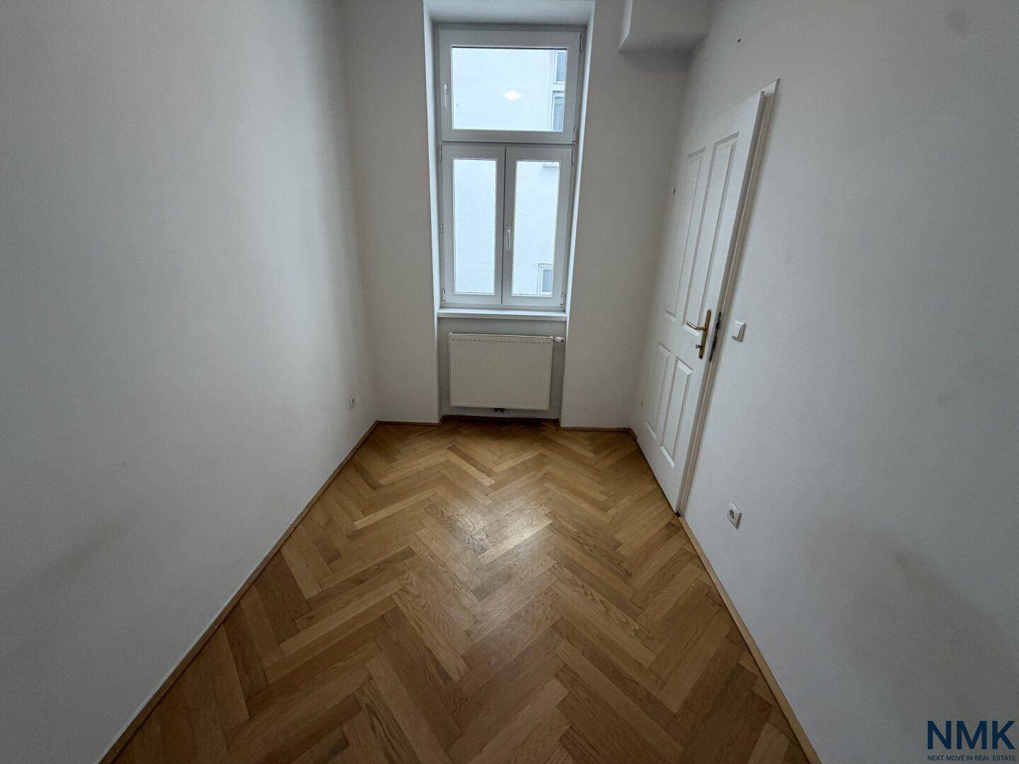 Helles Zimmer mit Fischgrätparkett und einem hohen Fenster.
