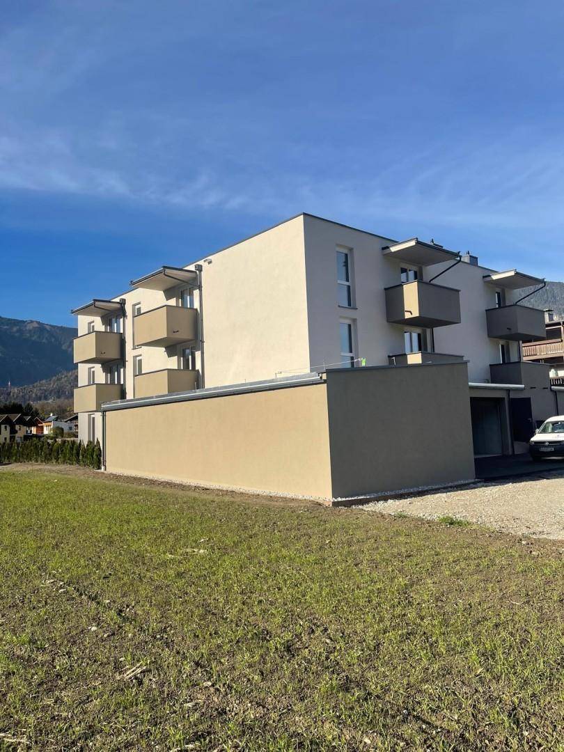Neubau-Wohngebäude mit Balkonen und angrenzender Grünfläche vor Bergkulisse.