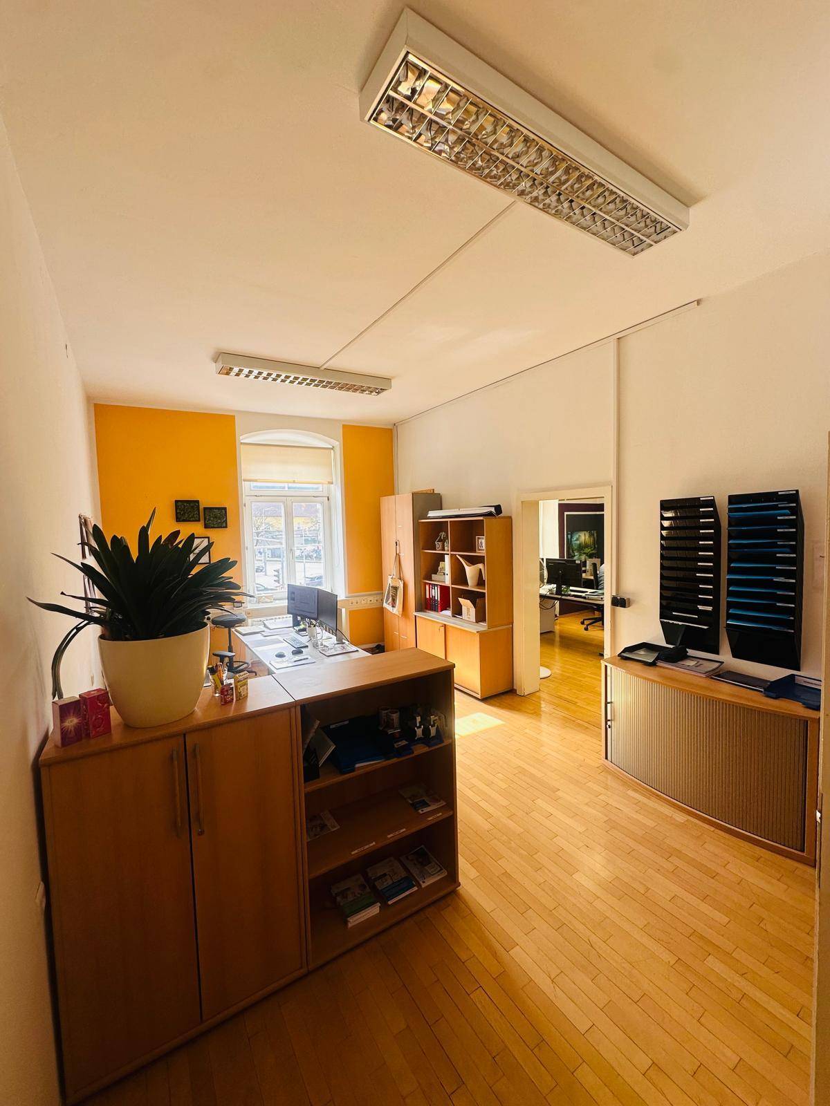 Helles Büro mit Holzfußboden, Schreibtisch und Regalen, durch eine Tür ist ein weiterer Raum sichtbar.