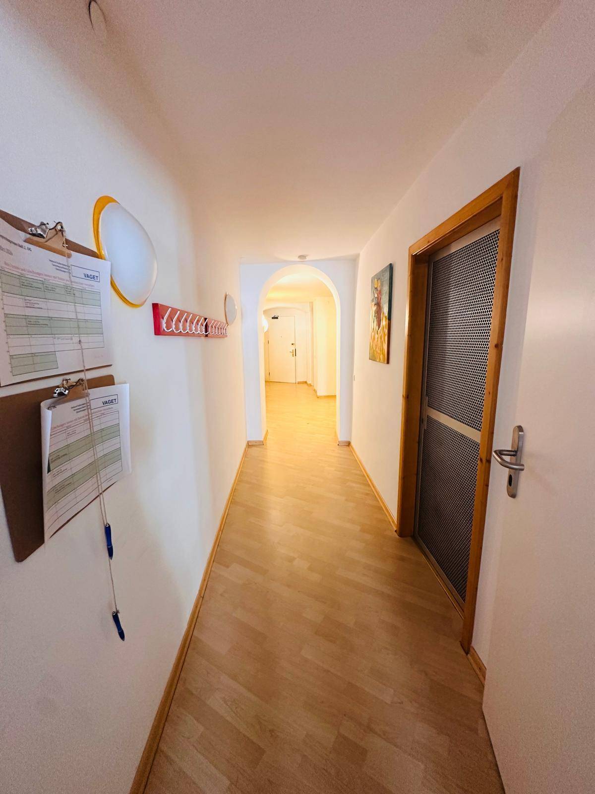 Heller Flur mit Holzfußboden, weißen Wänden und einem Bogen am Ende, der zu weiteren Räumen führt.