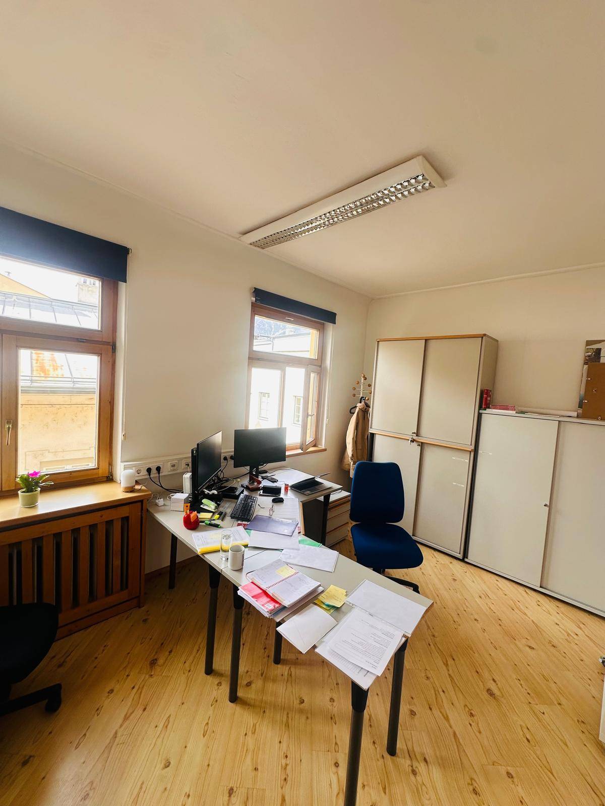 Einzelbüro mit Schreibtisch, Bürostuhl, Aktenschränken und zwei Fenstern, die viel Tageslicht hereinlassen.