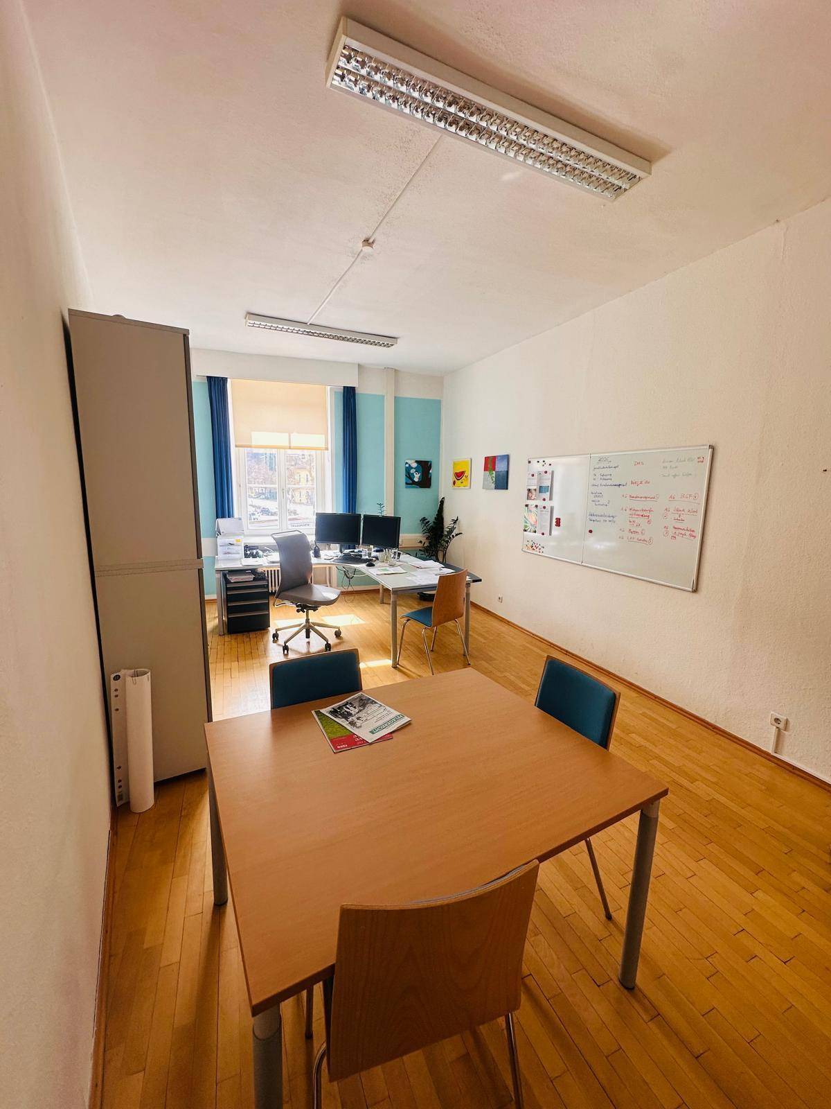 Helles Büro mit Holzfußboden, mehreren Schreibtischen und einem Besprechungstisch in der Mitte.