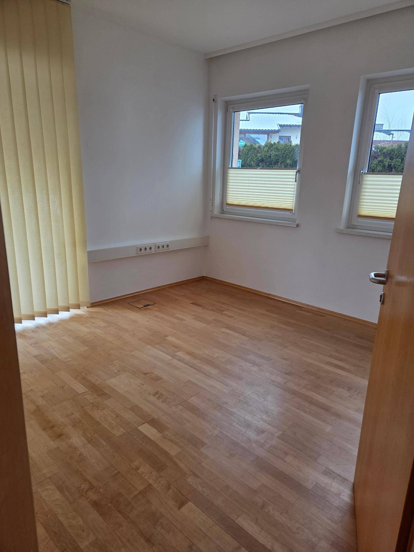 Helles Büro mit Holzparkettboden, zwei Fenstern und vertikalen Jalousien für Privatsphäre.