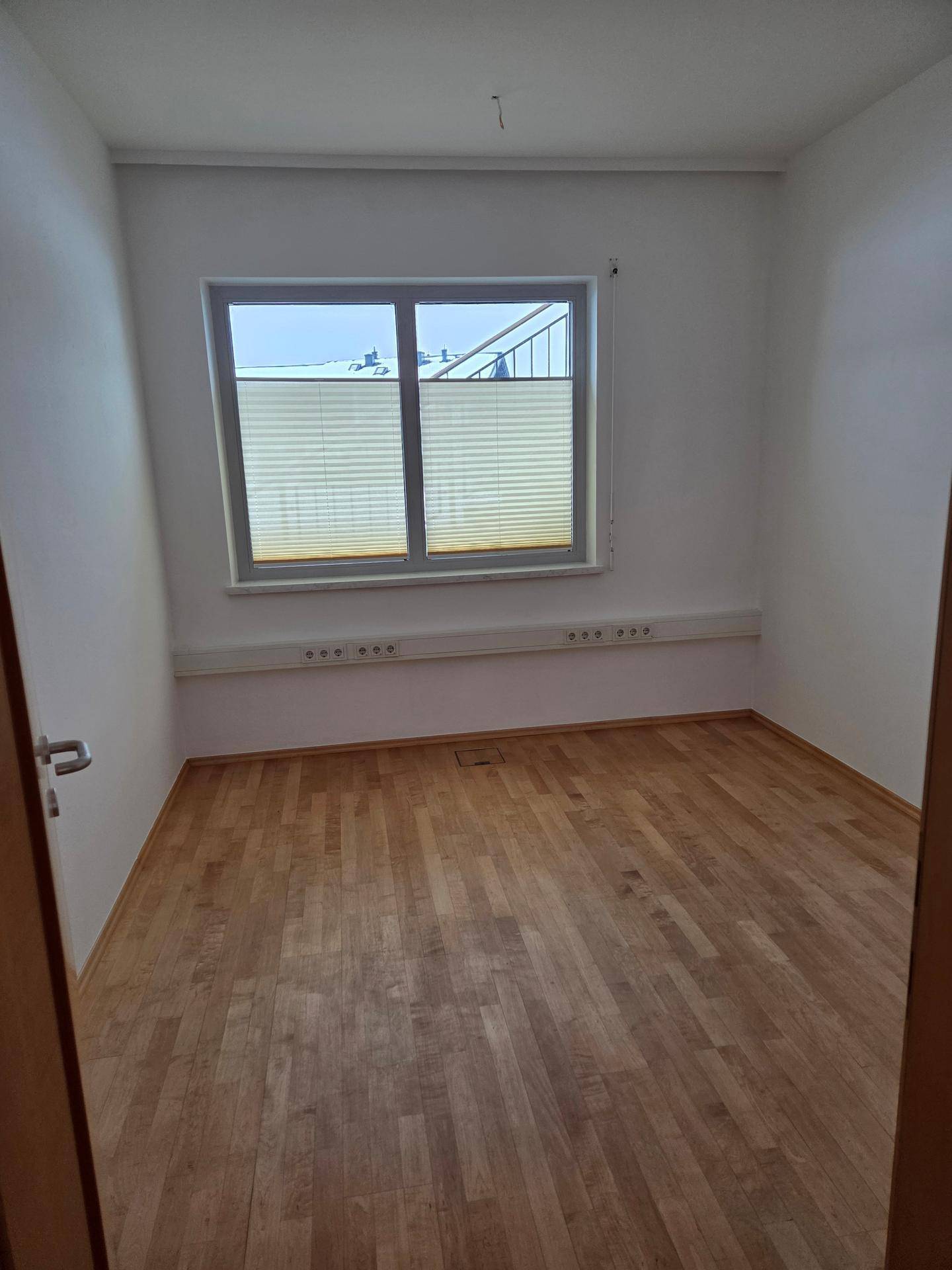 Kleineres Büro mit Holzboden und einem großen Fenster, das den Raum gut beleuchtet.