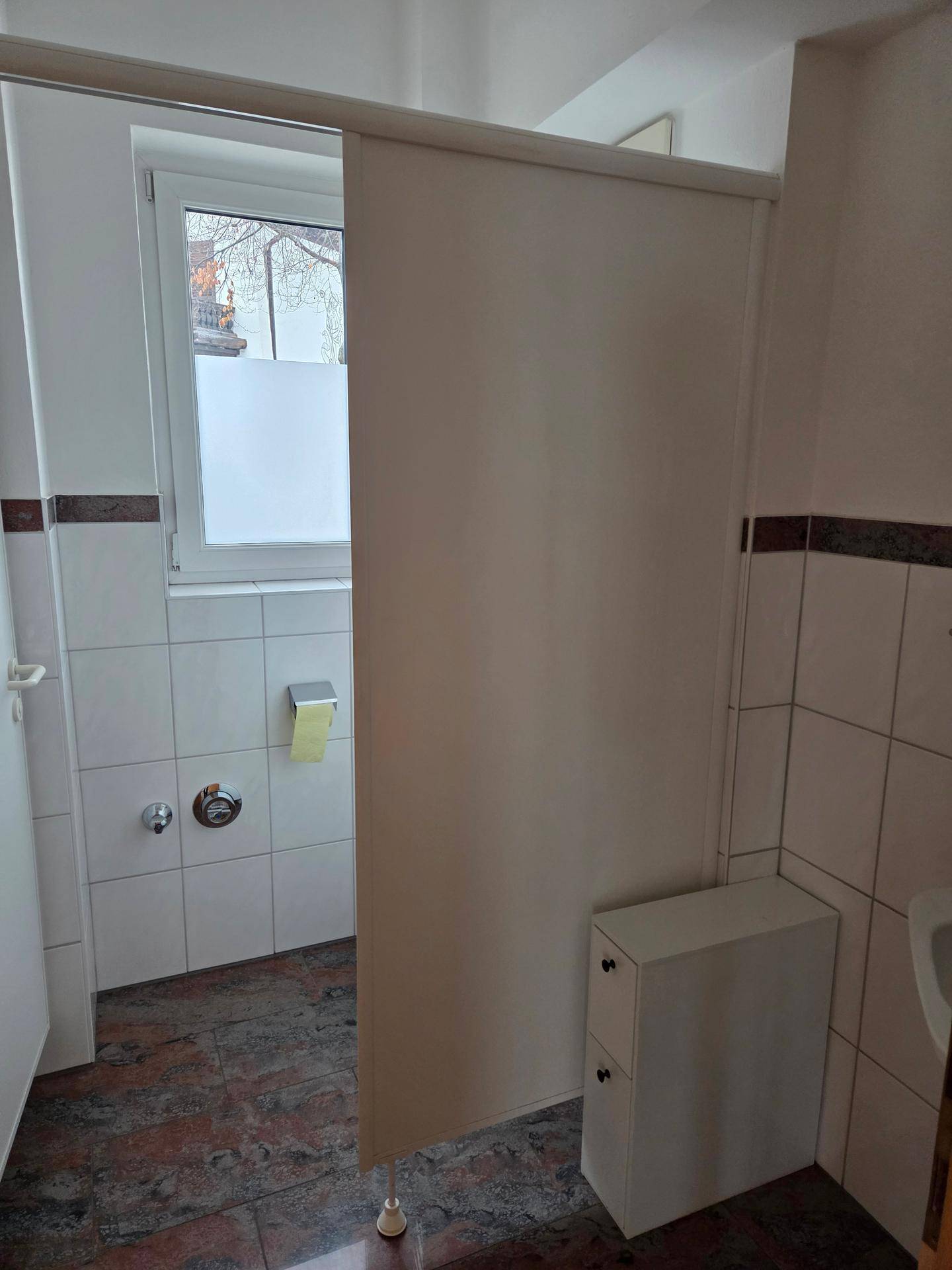 Badezimmer mit Fenster, weißen Fliesen und einem Duschvorhang, der den Duschbereich abtrennt.