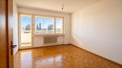 Leerer Raum mit Parkettboden und großem Fenster, das Zugang zu einem Balkon bietet.