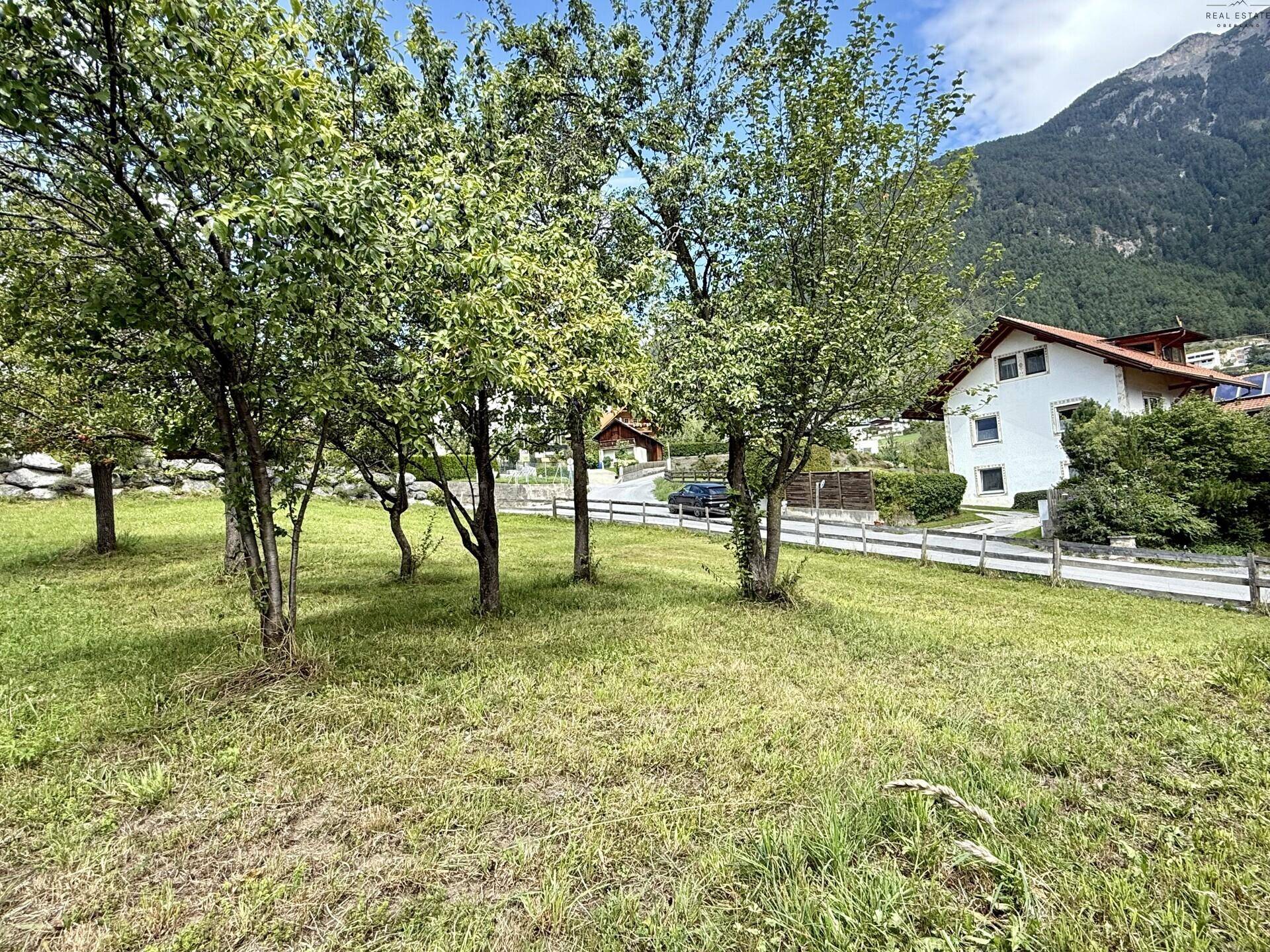 Grüne Wiese mit Obstbäumen und einem Haus im Hintergrund, umgeben von Bergen.