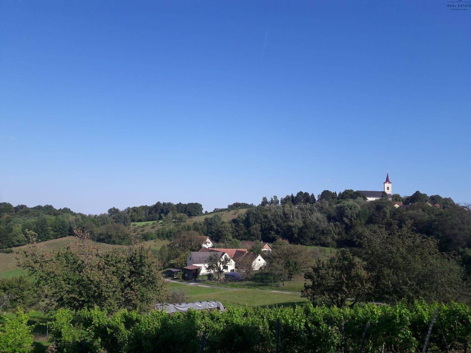 Panoramablick auf ein Dorf mit Kirche, umgeben von grünen Hügeln und Weinbergen unter blauem Himmel, der Weite vermittelt.
