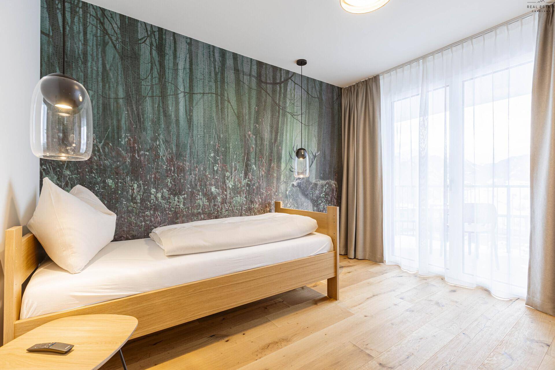 Helles Schlafzimmer mit Einzelbett, Holzboden und Waldmotiv-Tapete sowie Balkonzugang.