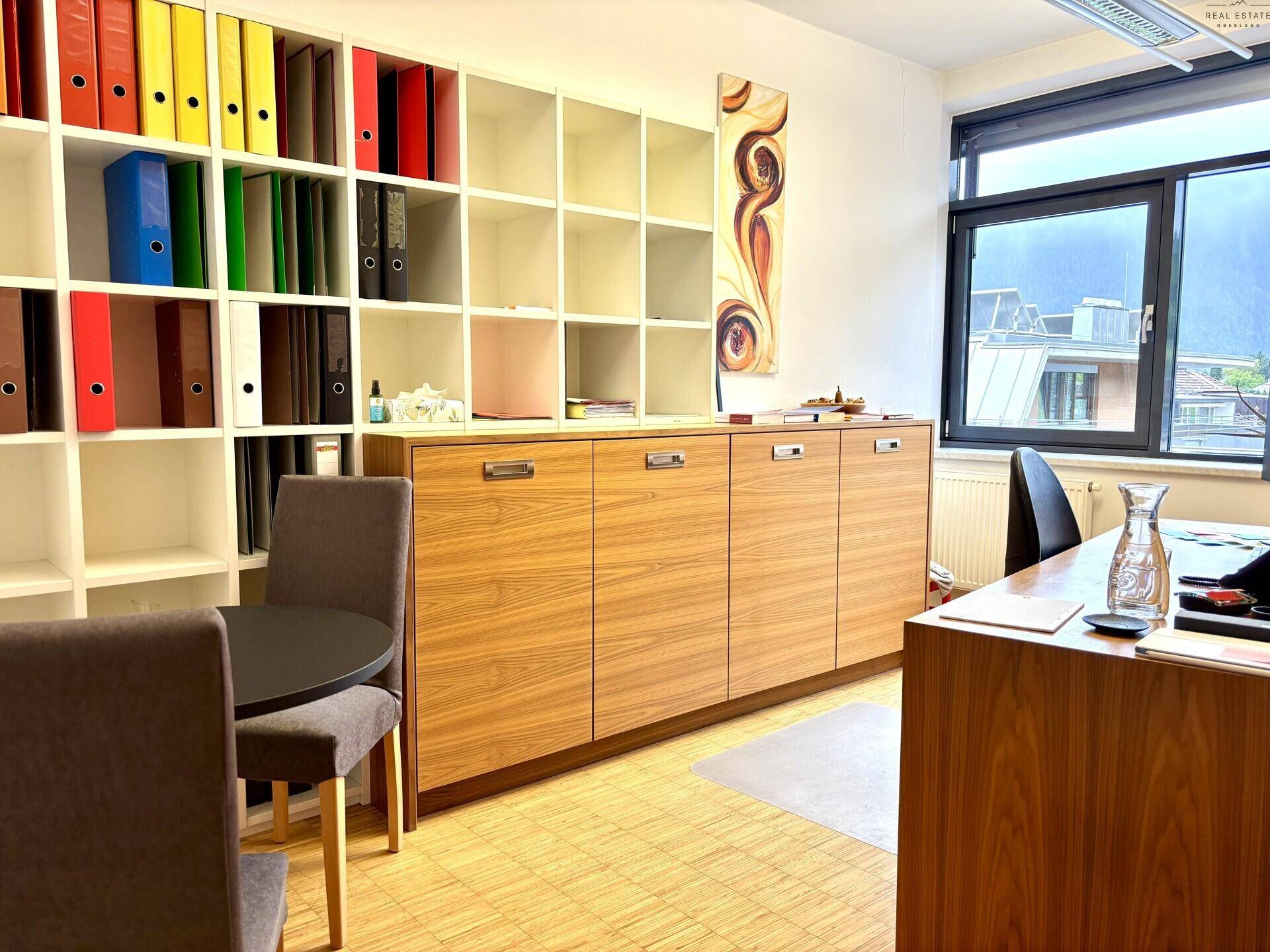 Bürobereich mit Holzschränken, weißen Regalen voller Ordner und einem kleinen runden Tisch.