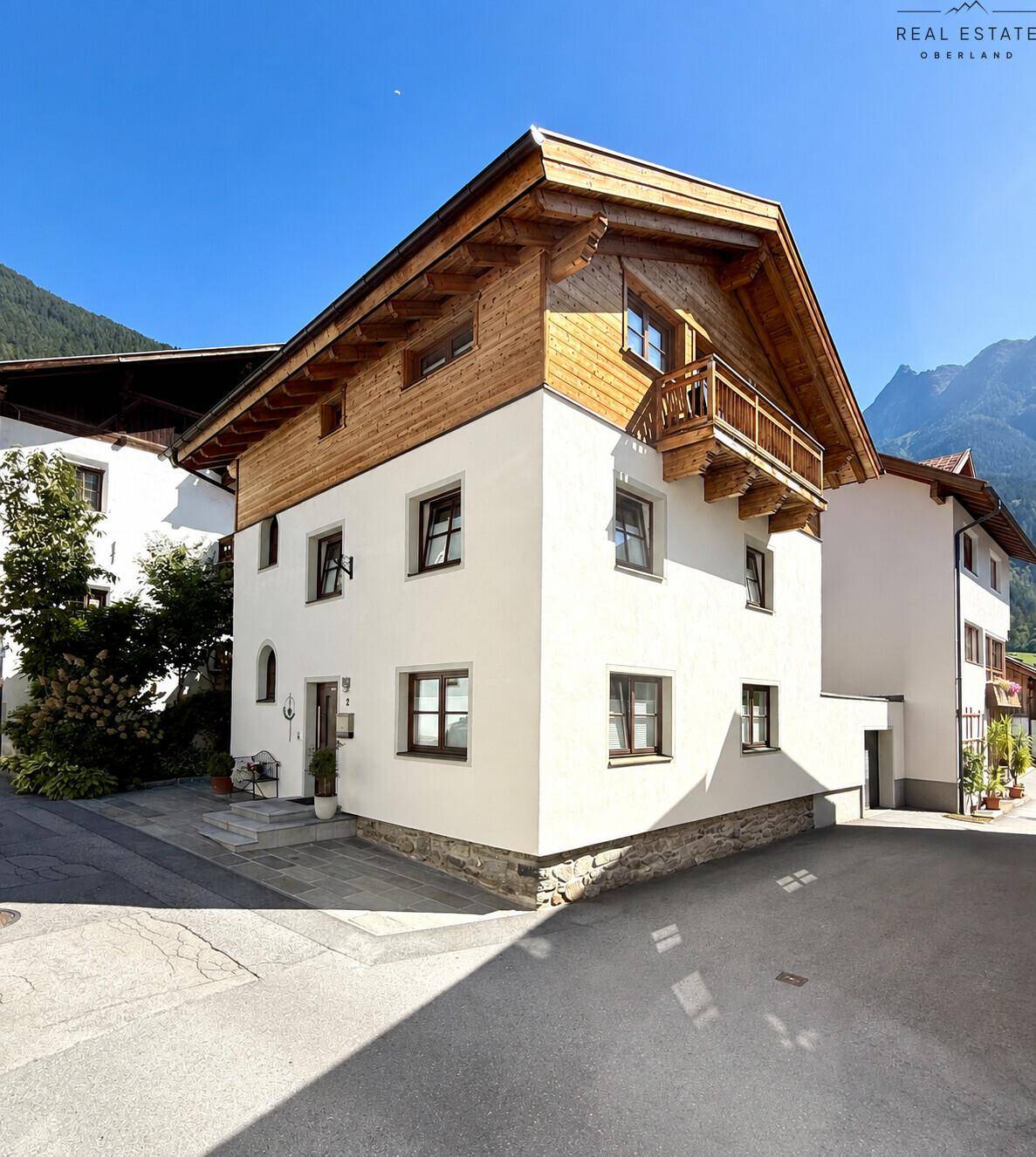 Charmantes Haus mit Holzbalkon und weißer Fassade vor alpiner Bergkulisse.