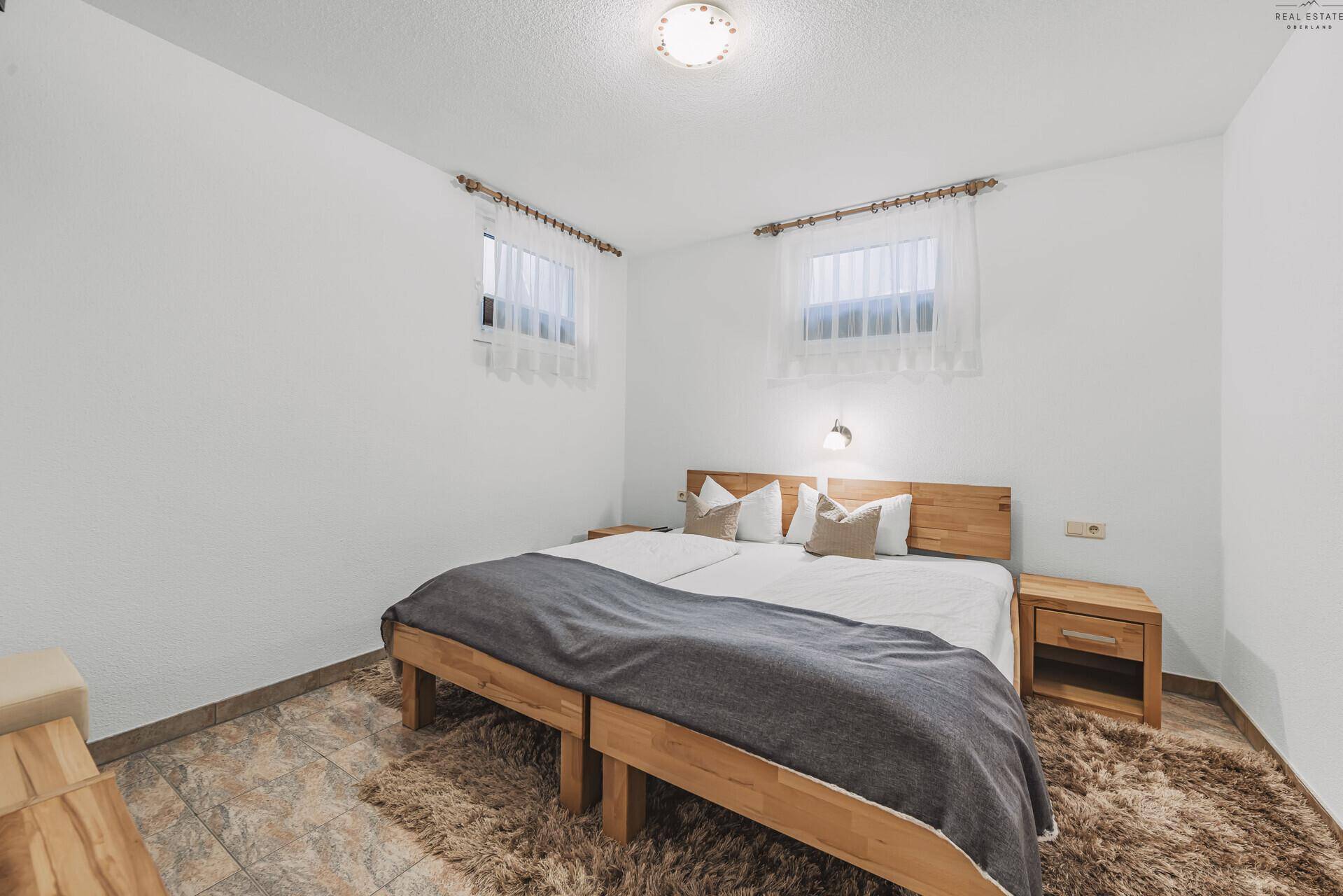 Gemütliches Schlafzimmer mit Doppelbett aus Holz, Fliesenboden und zwei Fenstern.