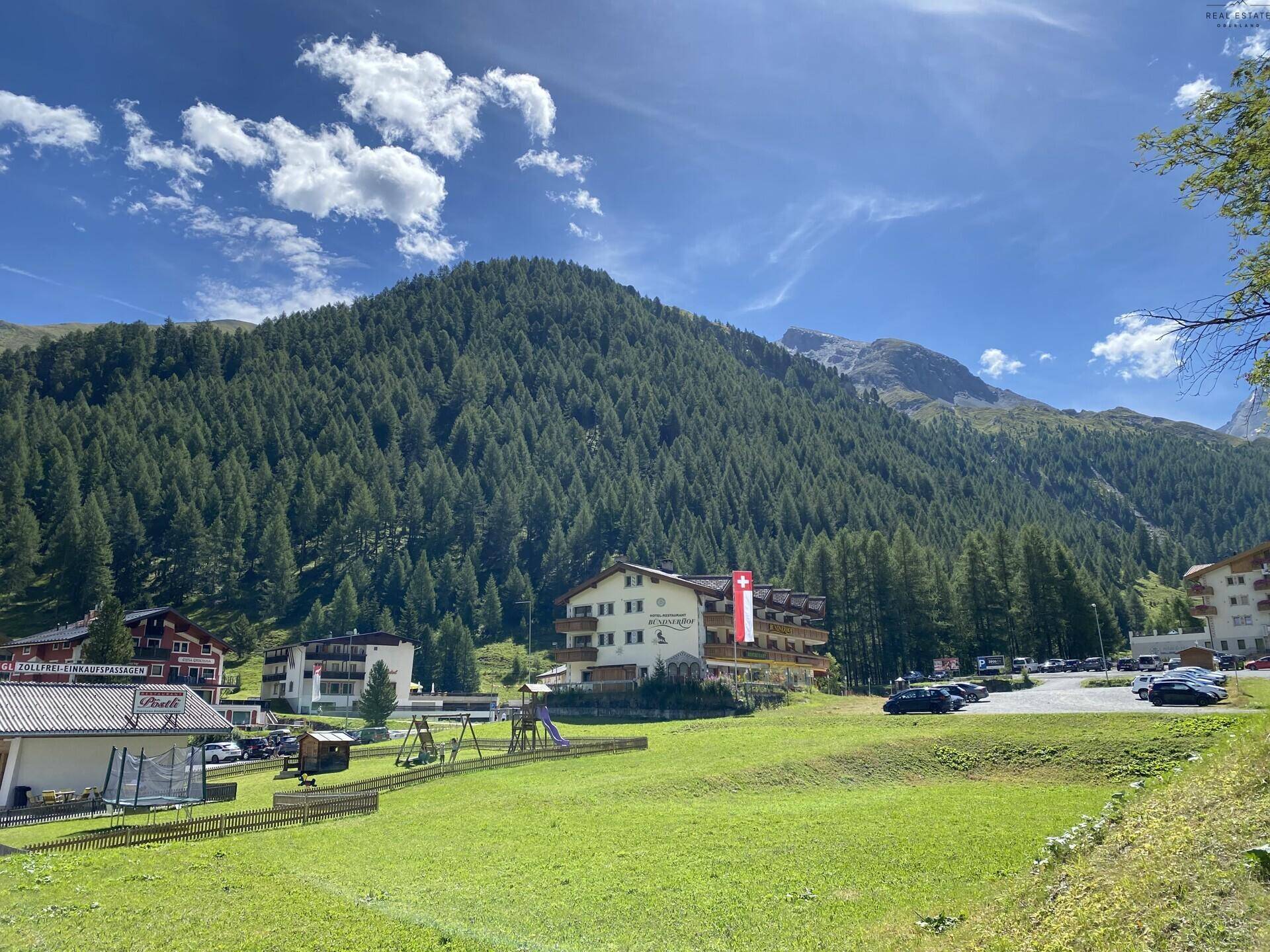 Hotelgebäude in einer malerischen Berglandschaft mit Wald, Wiesen und Spielplatz.