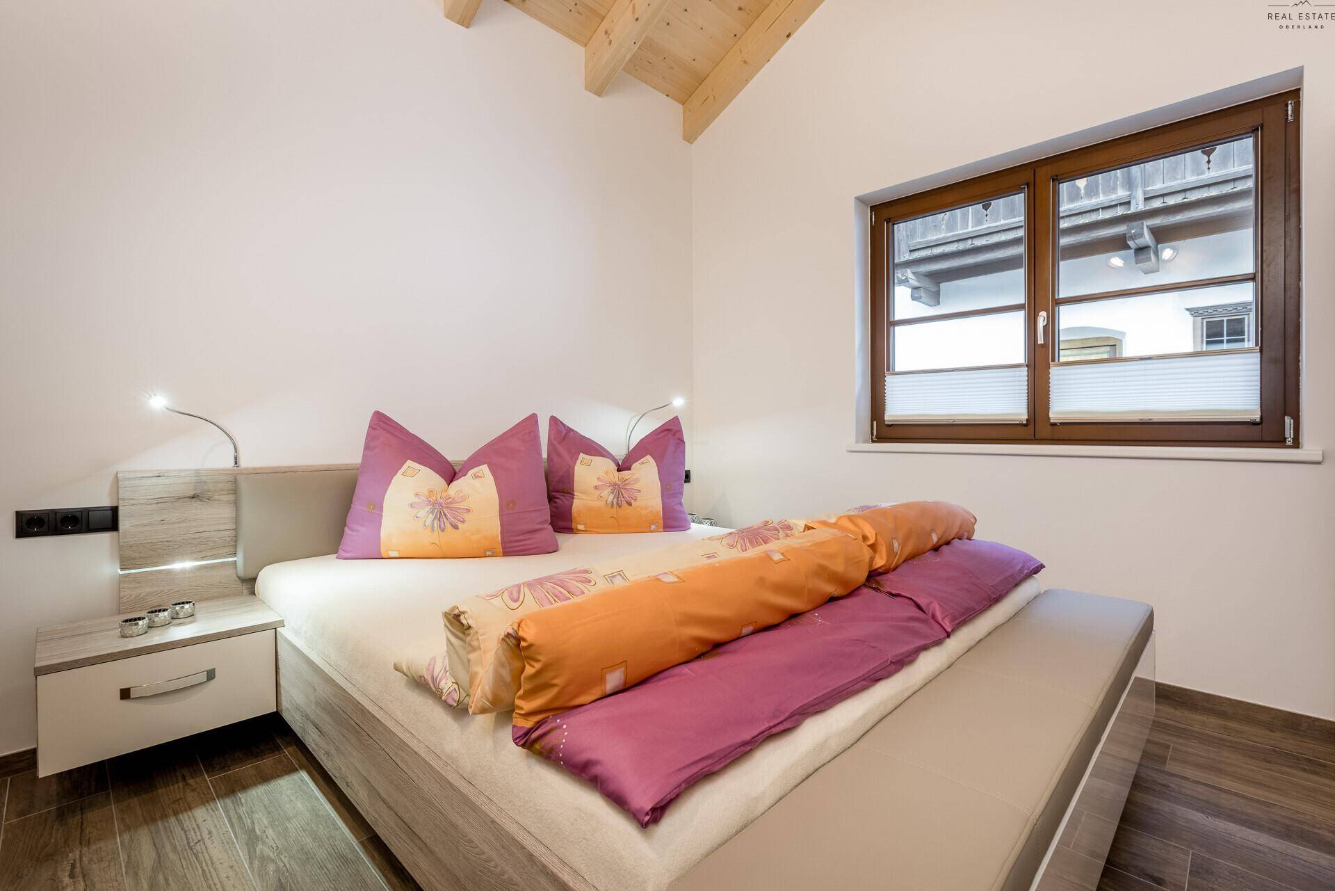 Helles Schlafzimmer mit Doppelbett, Holzbalkendecke und Fenster mit Blick nach draußen.