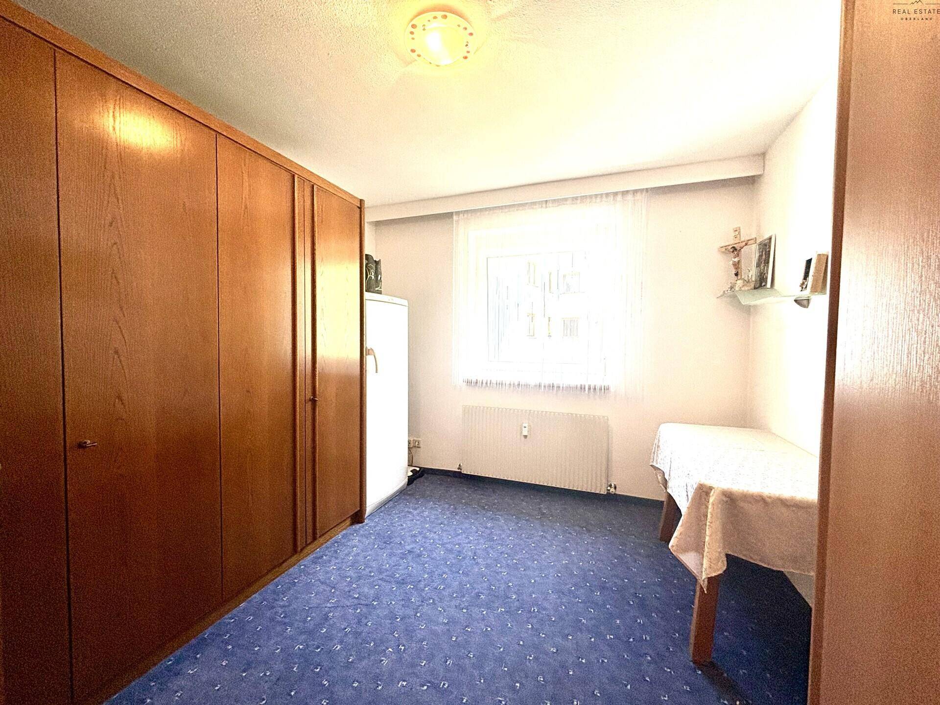 Schlafzimmer mit großem Holzschrank, blauem Teppichboden und einem Fenster, das viel Licht hereinlässt.