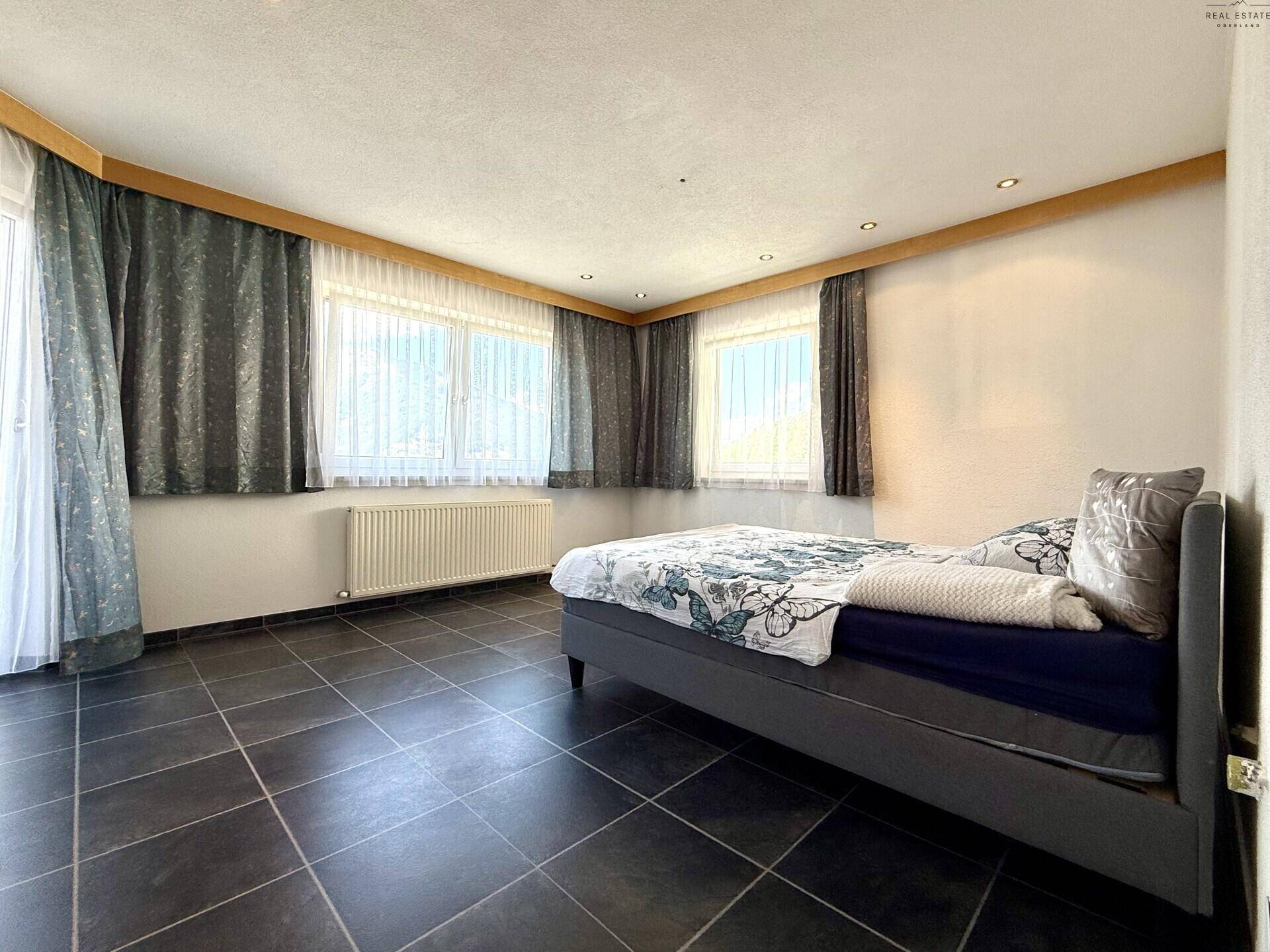 Schlafzimmer mit dunklem Fliesenboden, Doppelbett und großen Fenstern, die viel Licht hereinlassen.