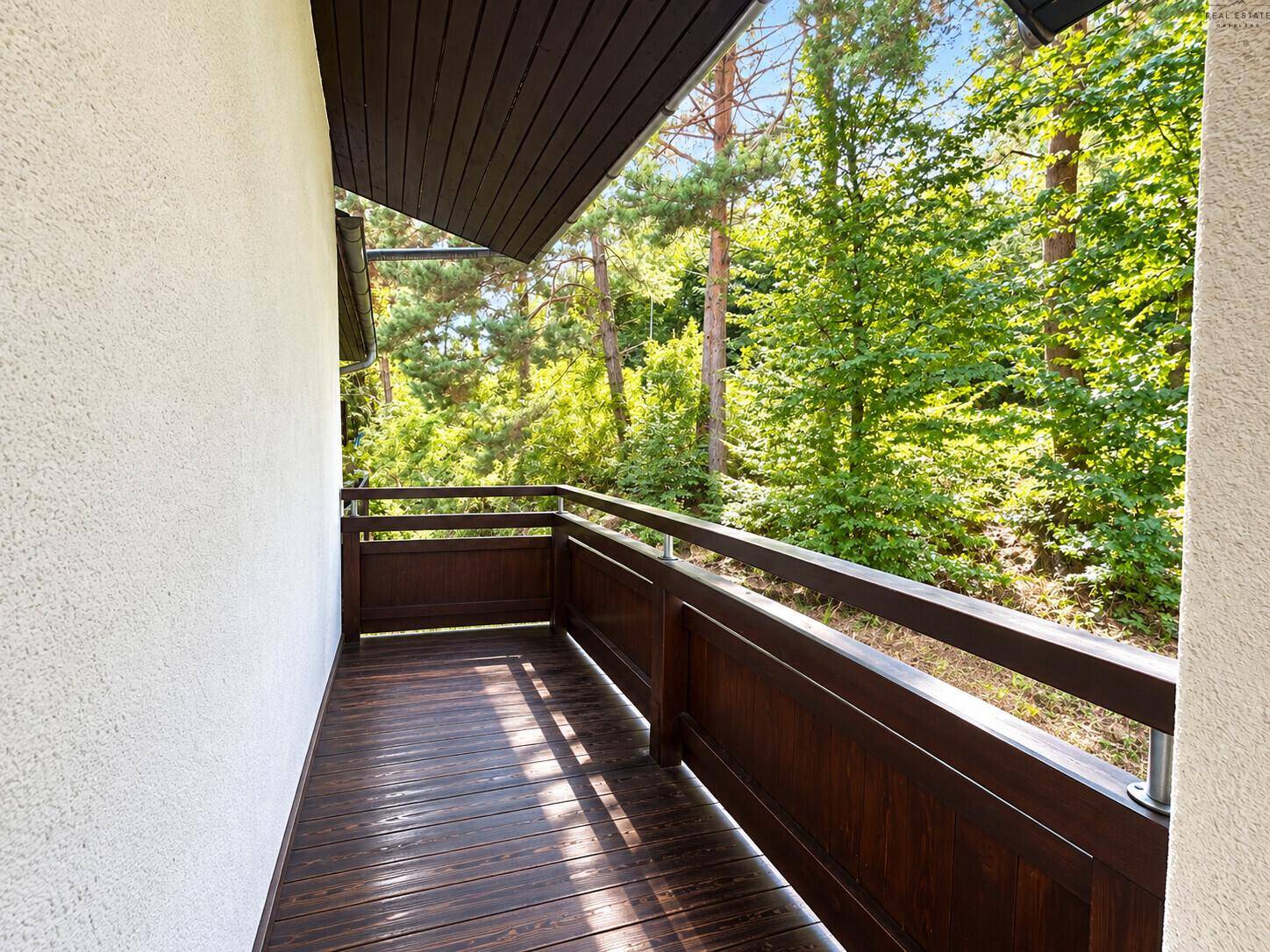Heller Balkon mit Holzboden und Blick ins Grüne, umgeben von Bäumen.