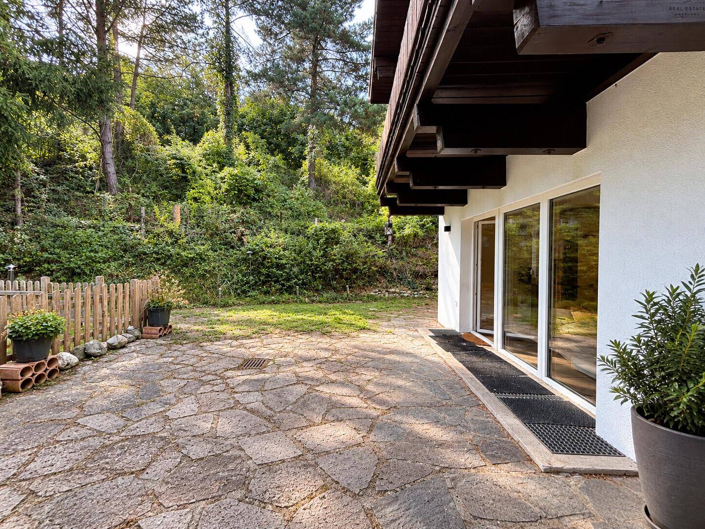 Gepflasterte Terrasse am Haus mit Holzzaun und üppiger grüner Bepflanzung im Hintergrund.
