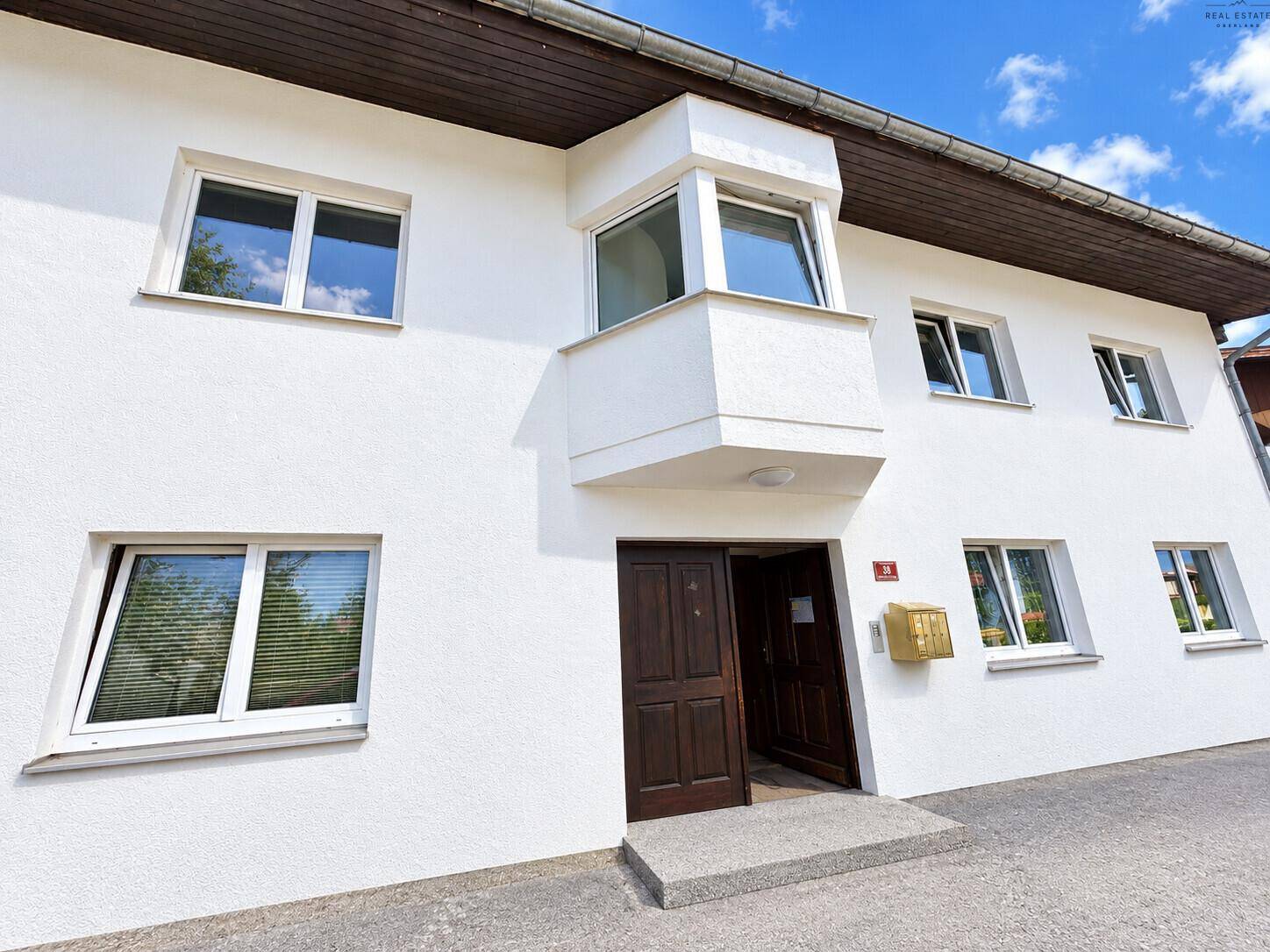 Eingangsbereich eines weißen Hauses mit Holztür und einem kleinen Erker im Obergeschoss.