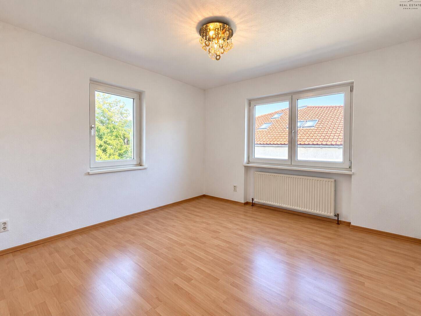 Leeres Schlafzimmer mit Laminatboden, zwei Fenstern und Deckenleuchte.