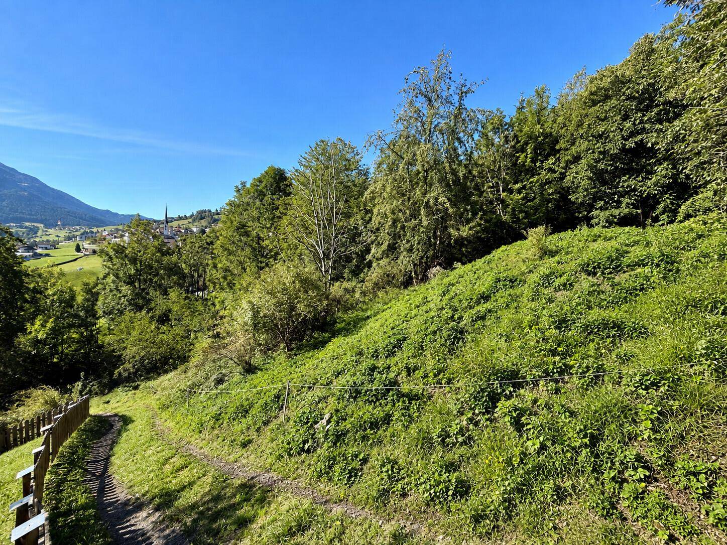 Grüner Hang mit einem schmalen Pfad und Blick auf ein Dorf in der Ferne.