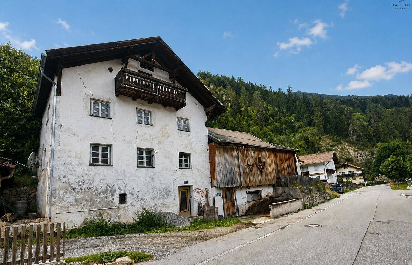 Historisches Bauernhaus mit Holzbalkon und angrenzendem Holzgebäude in ländlicher Umgebung.
