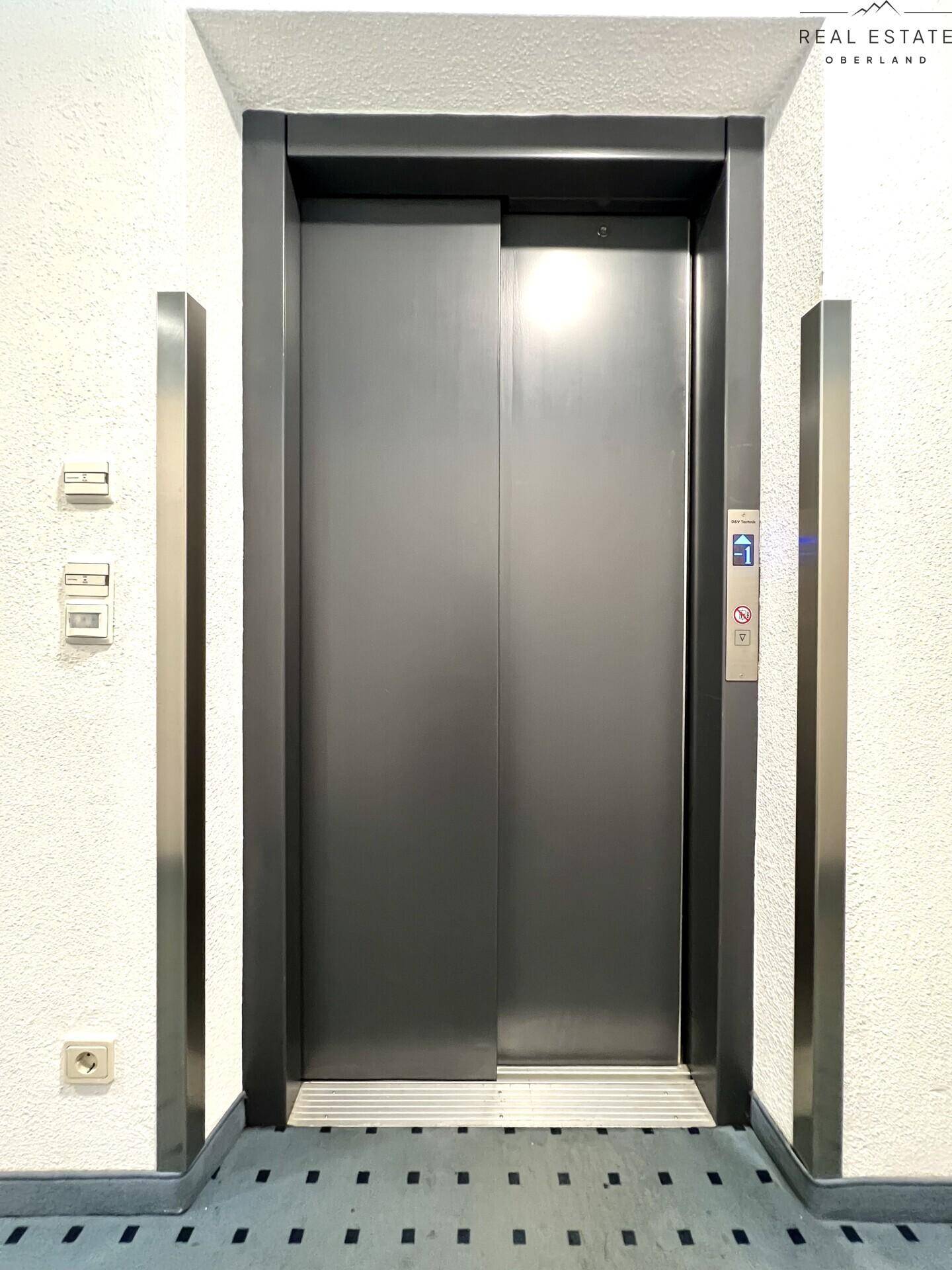 Moderner Aufzug mit grauen Türen und Edelstahlverkleidung in einem gut beleuchteten Flur.