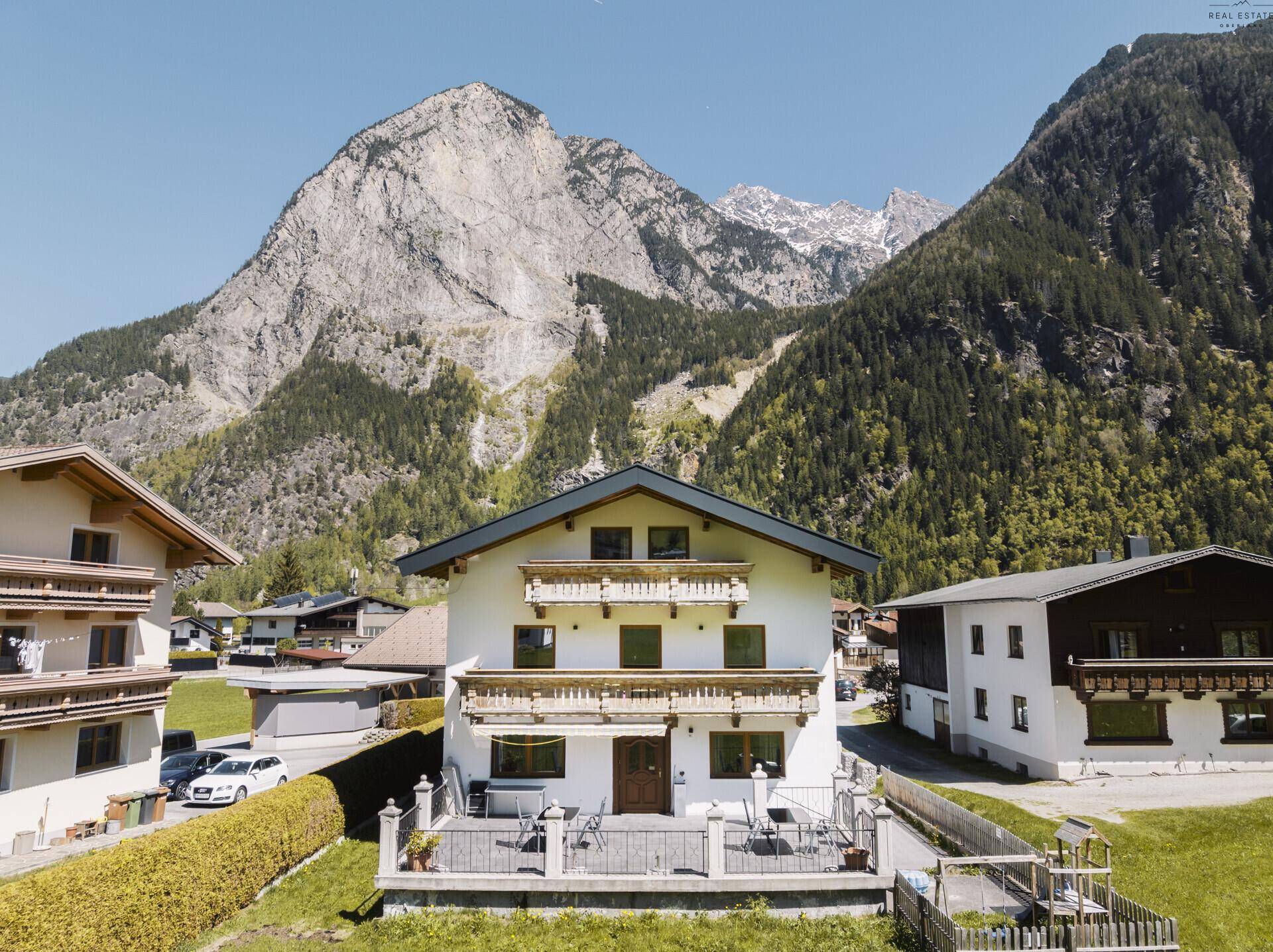 Traditionelles Haus mit Balkonen und Terrasse, umgeben von majestätischen Bergen und grüner Landschaft.