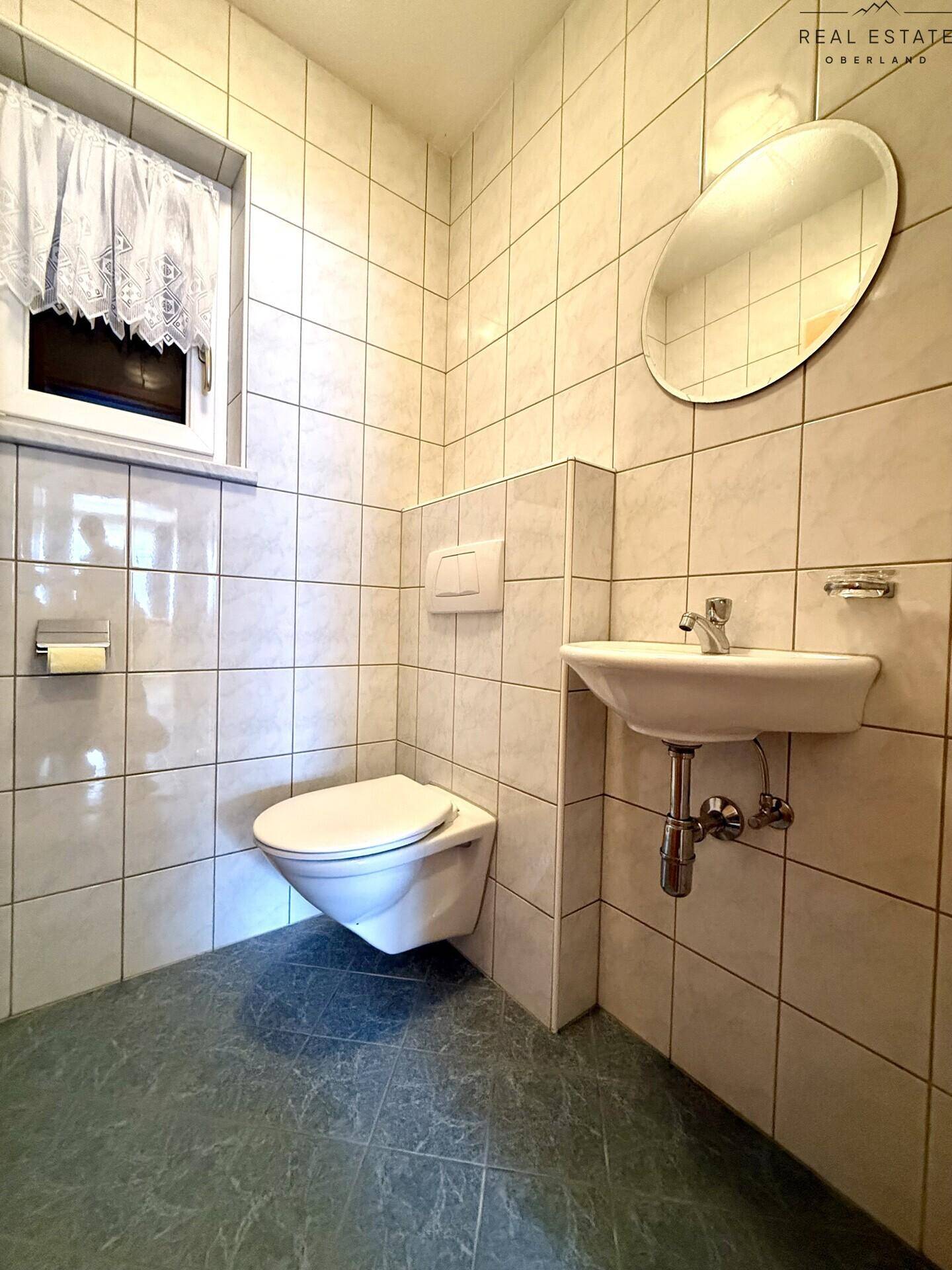 Kleines Badezimmer mit WC, Waschbecken, Spiegel und Fliesenboden in hellen Farben.