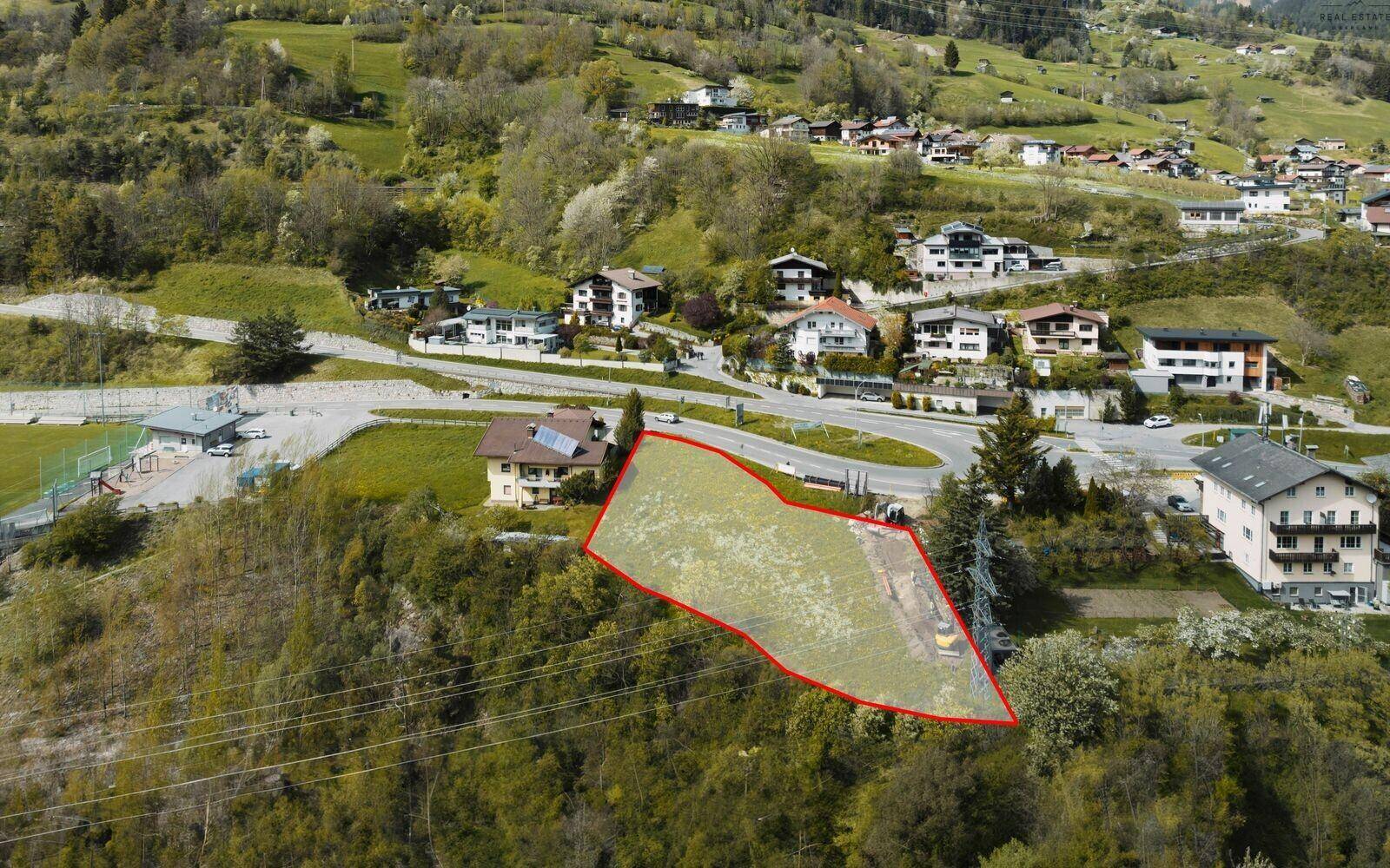 Luftaufnahme eines Baugrundstücks mit roter Markierung, umgeben von Wohnhäusern und Bergen.