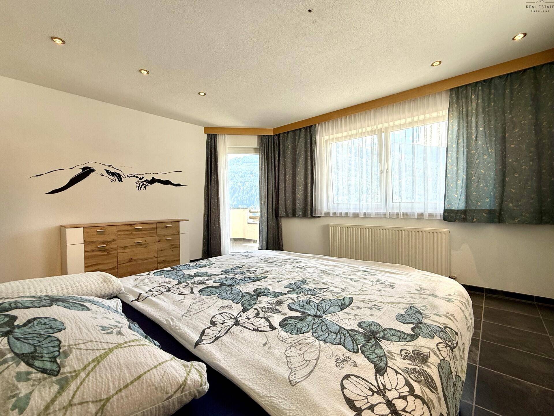 Schlafzimmer mit Doppelbett, dunklem Fliesenboden und Fenstern mit Bergblick und viel Tageslicht.