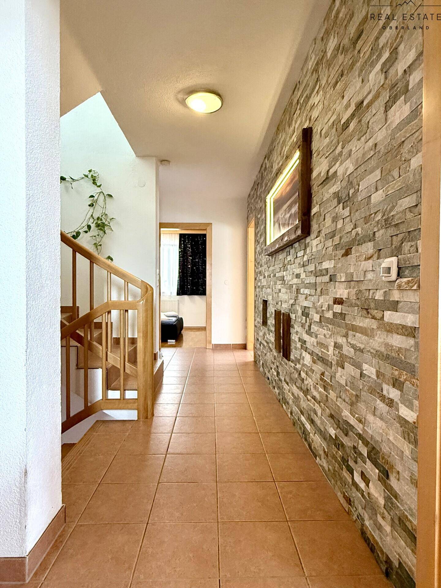 Heller Flur mit Steinfurnierwand, Holztreppe und Terrakotta-Fliesenboden.