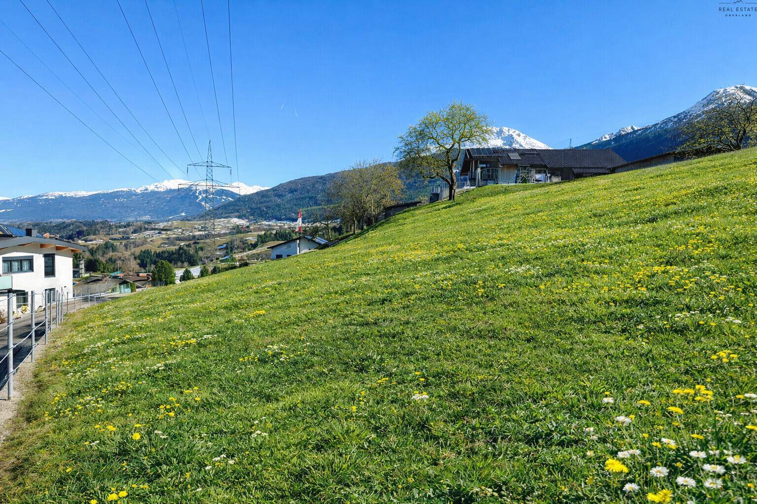 Sonnige Wiesenlandschaft mit vereinzelten Häusern und einem weiten Blick auf die Bergkette.