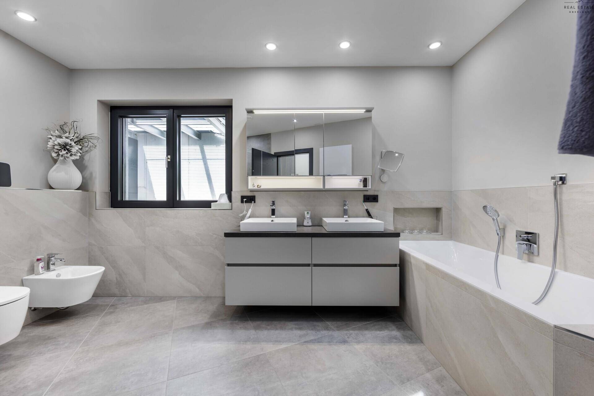 Modernes Badezimmer mit Doppelwaschbecken, Badewanne, Bidet und Fenster in hellen Grautönen.