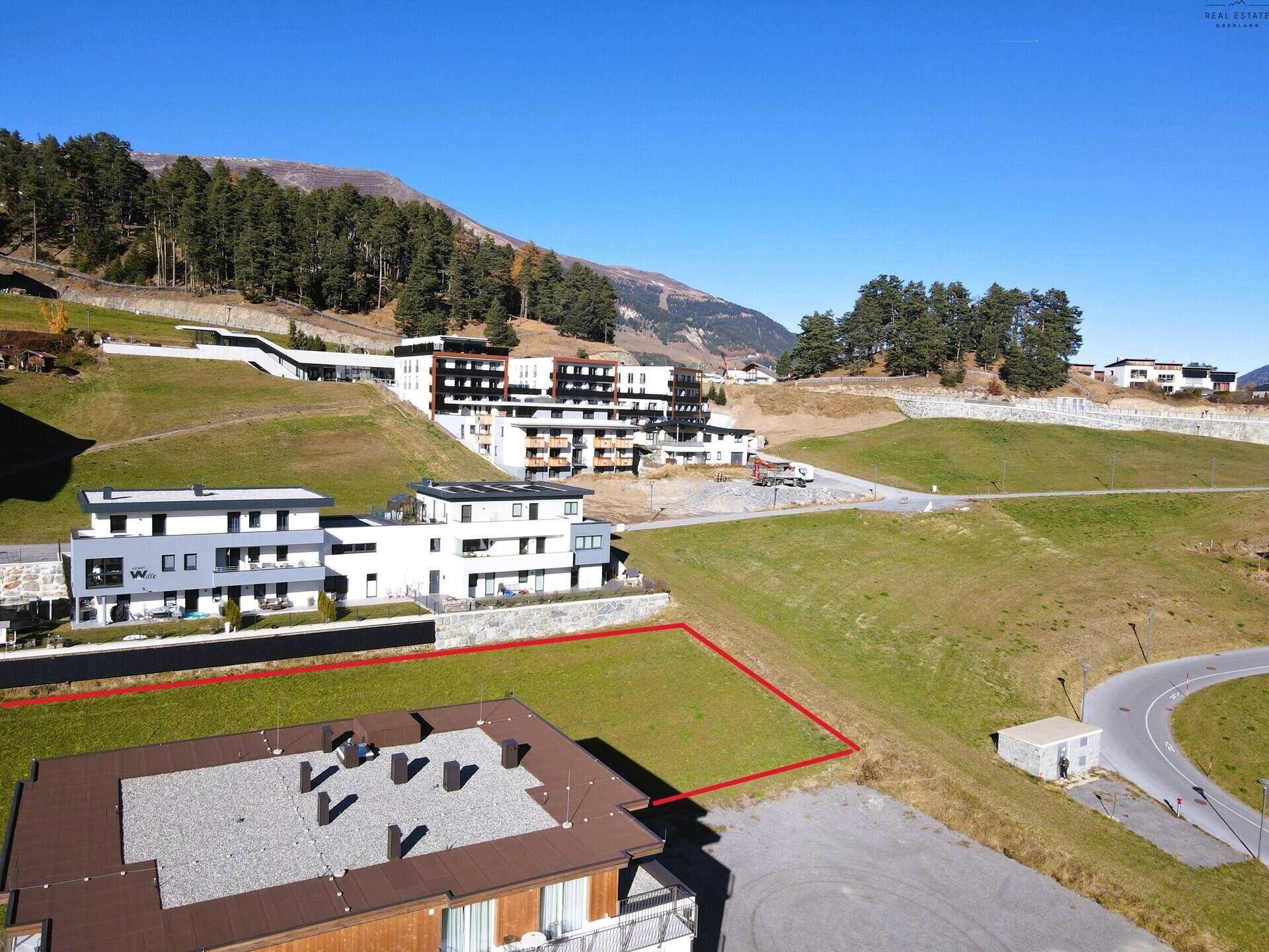 Luftaufnahme eines Baugrundstücks mit roter Markierung und Zufahrtsstraße in alpiner Umgebung.