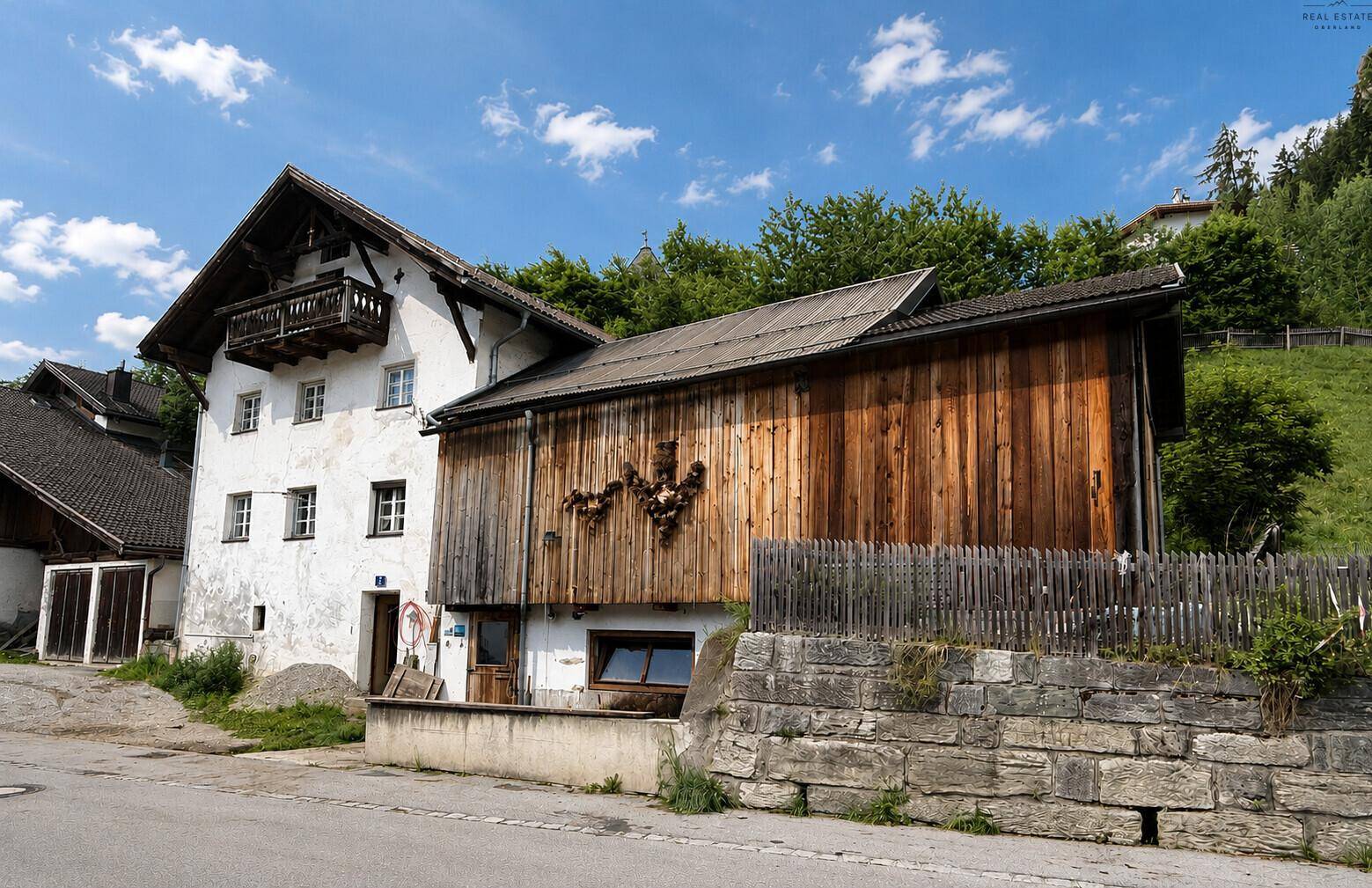 Altes Bauernhaus mit weißer Fassade und rustikalem Holzanbau, umgeben von grüner Natur.