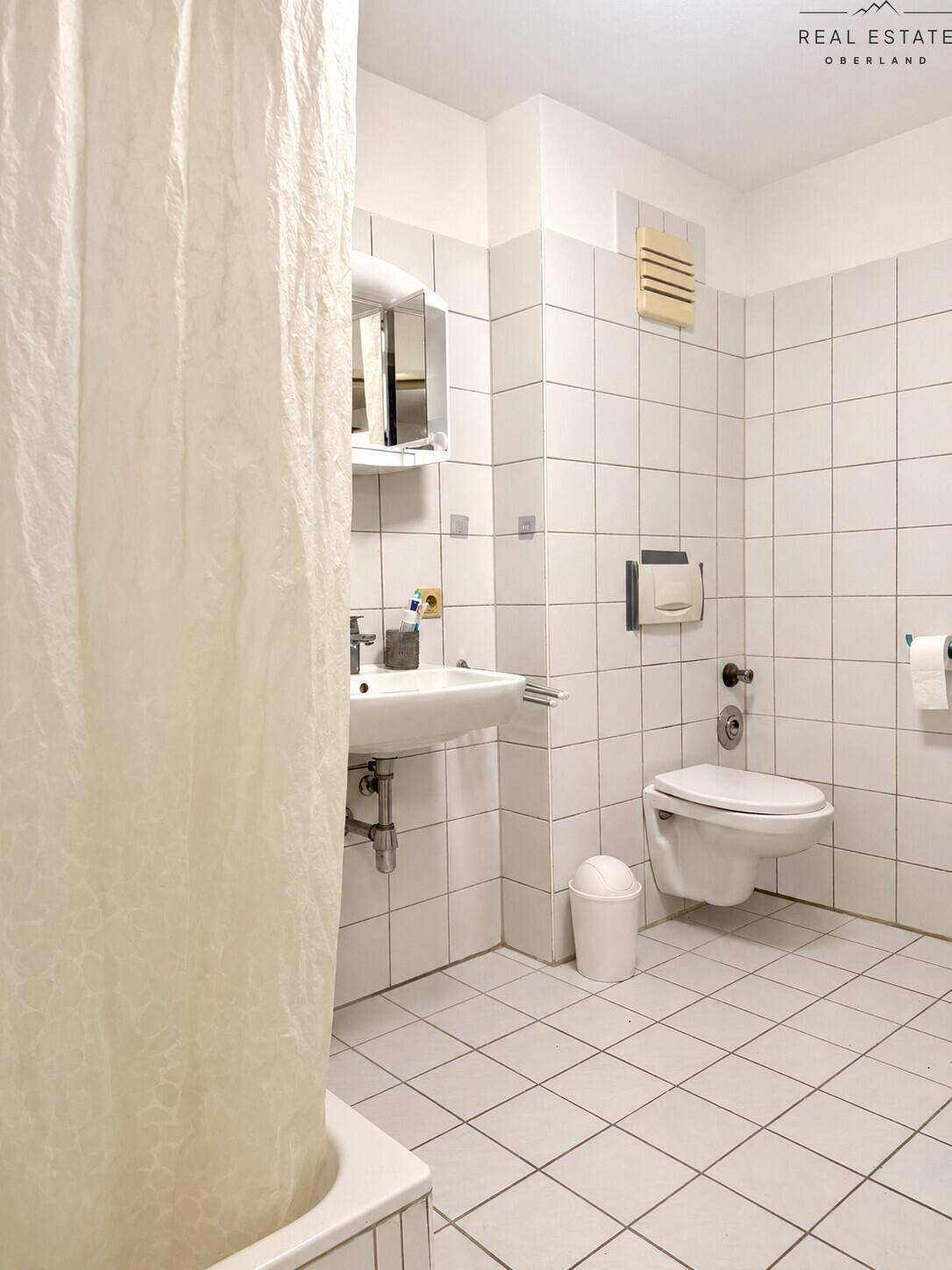 Helles Badezimmer mit weißen Fliesen, WC, Waschbecken und Duschvorhang.