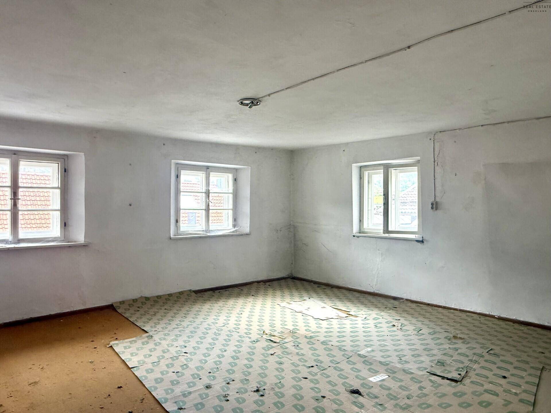 Leerer Raum mit weißen Wänden, mehreren Fenstern und altem Bodenbelag, renovierungsbedürftig.