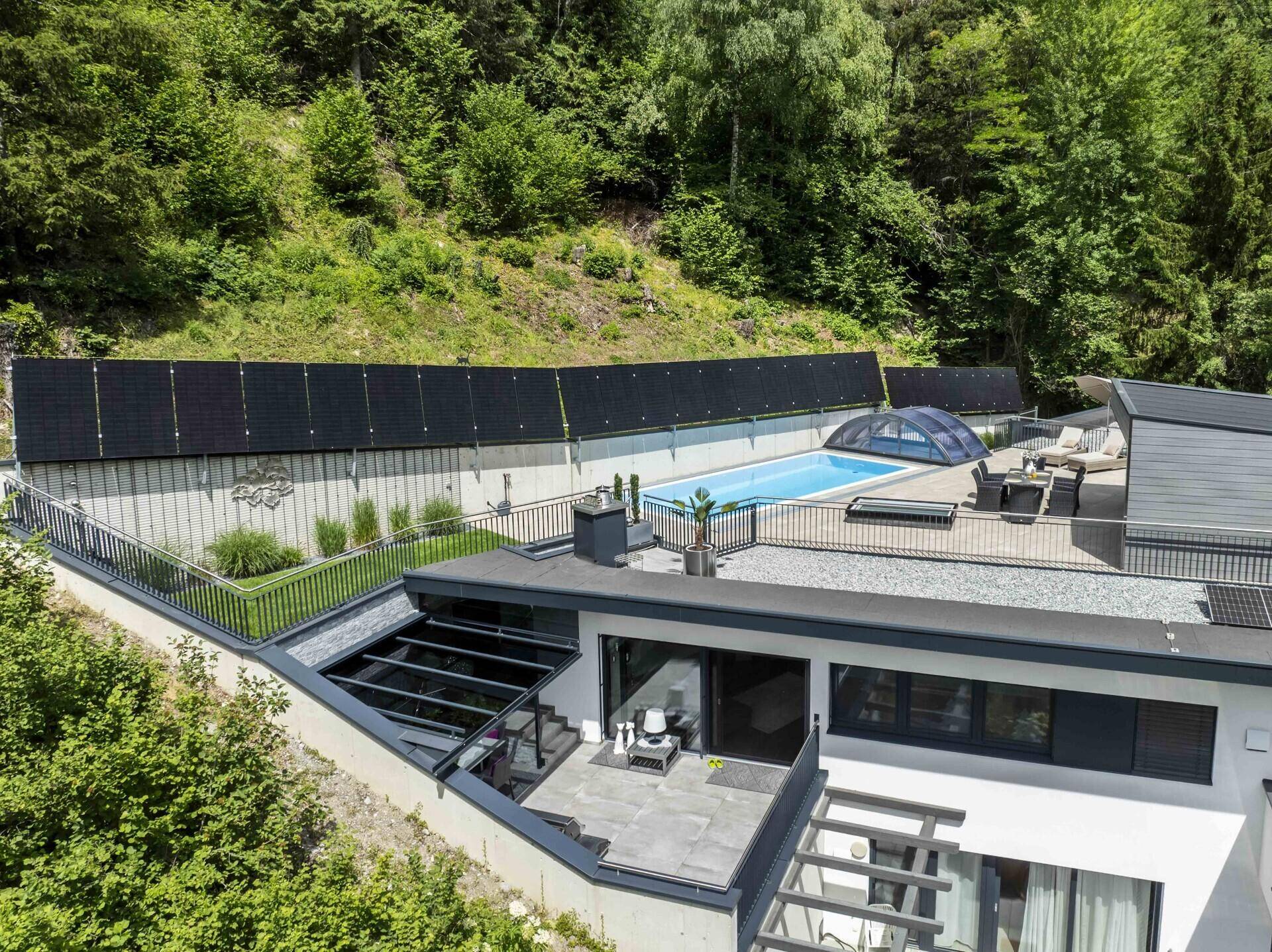 Weitläufige Außenansicht des Anwesens mit Poolbereich, Solaranlage und moderner Architektur vor bewaldetem Hang.
