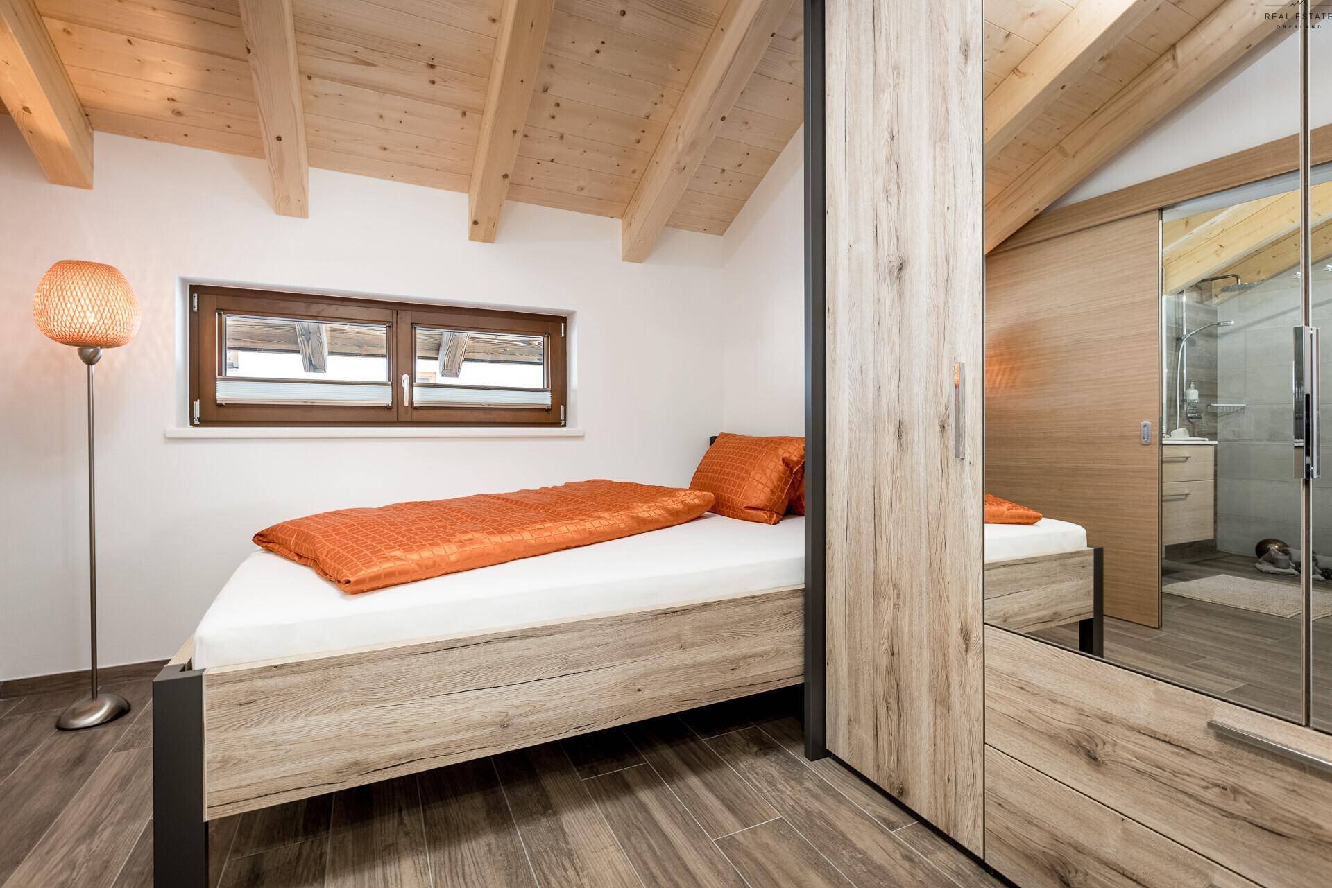 Helles Schlafzimmer mit Einzelbett, Holzbalkendecke und Blick auf das Badezimmer.