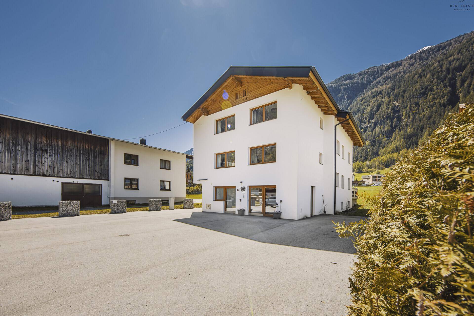 Weißes Wohnhaus mit Holzelementen und großem Vorplatz, umgeben von alpiner Landschaft.