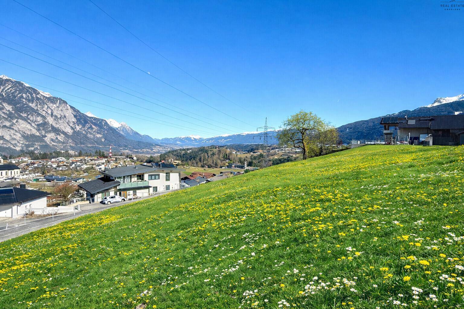 Weitläufige Landschaft mit grünen Wiesen, Häusern und schneebedeckten Bergen im Hintergrund.
