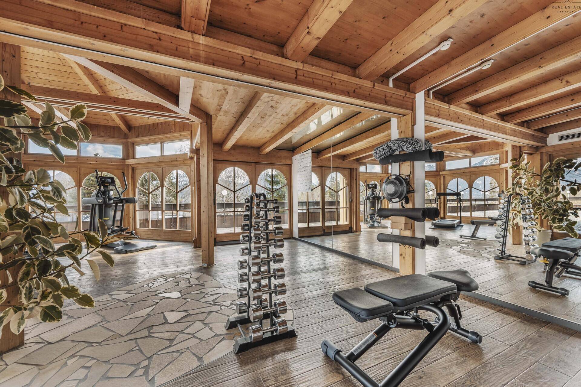 Gut ausgestatteter Fitnessraum mit verschiedenen Geräten, Holzbalken und großen Fenstern.