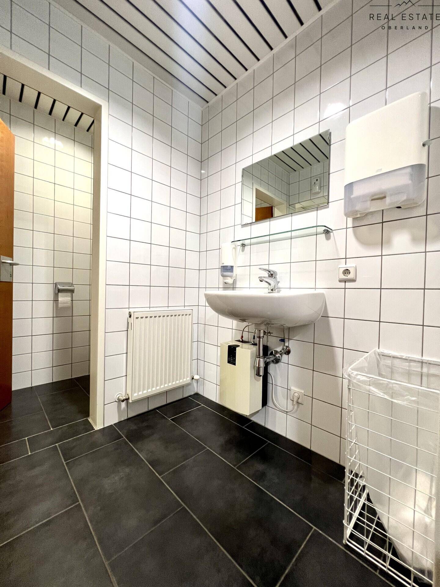 Funktionales Badezimmer mit weißen Fliesen, dunklem Boden und einem Waschbeckenbereich.