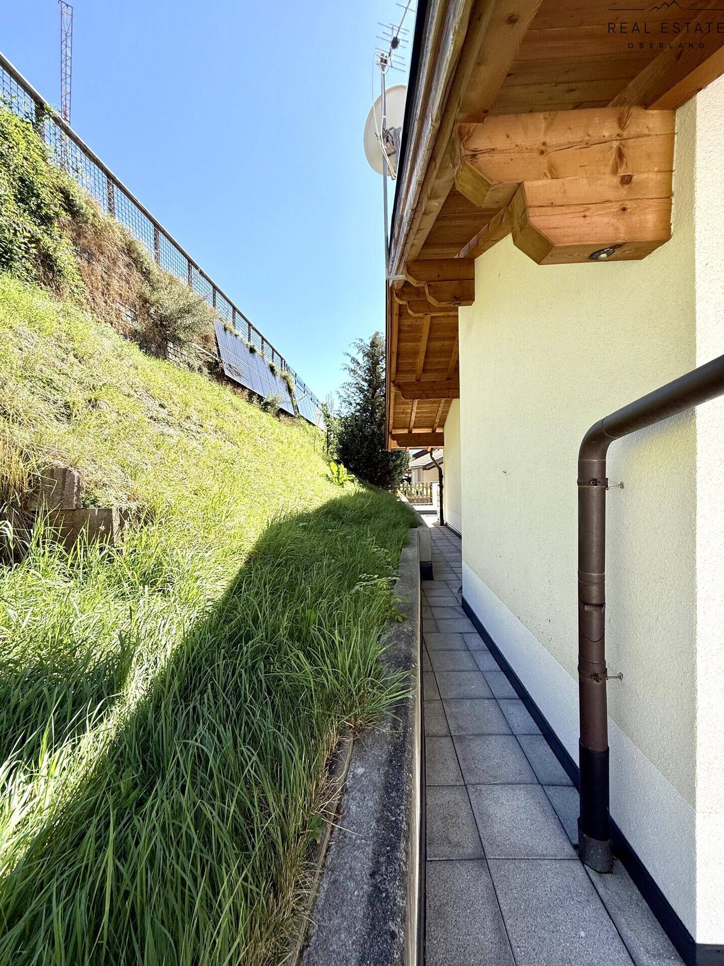 Schmaler gefliester Weg entlang der Hauswand mit angrenzendem begrüntem Hang.