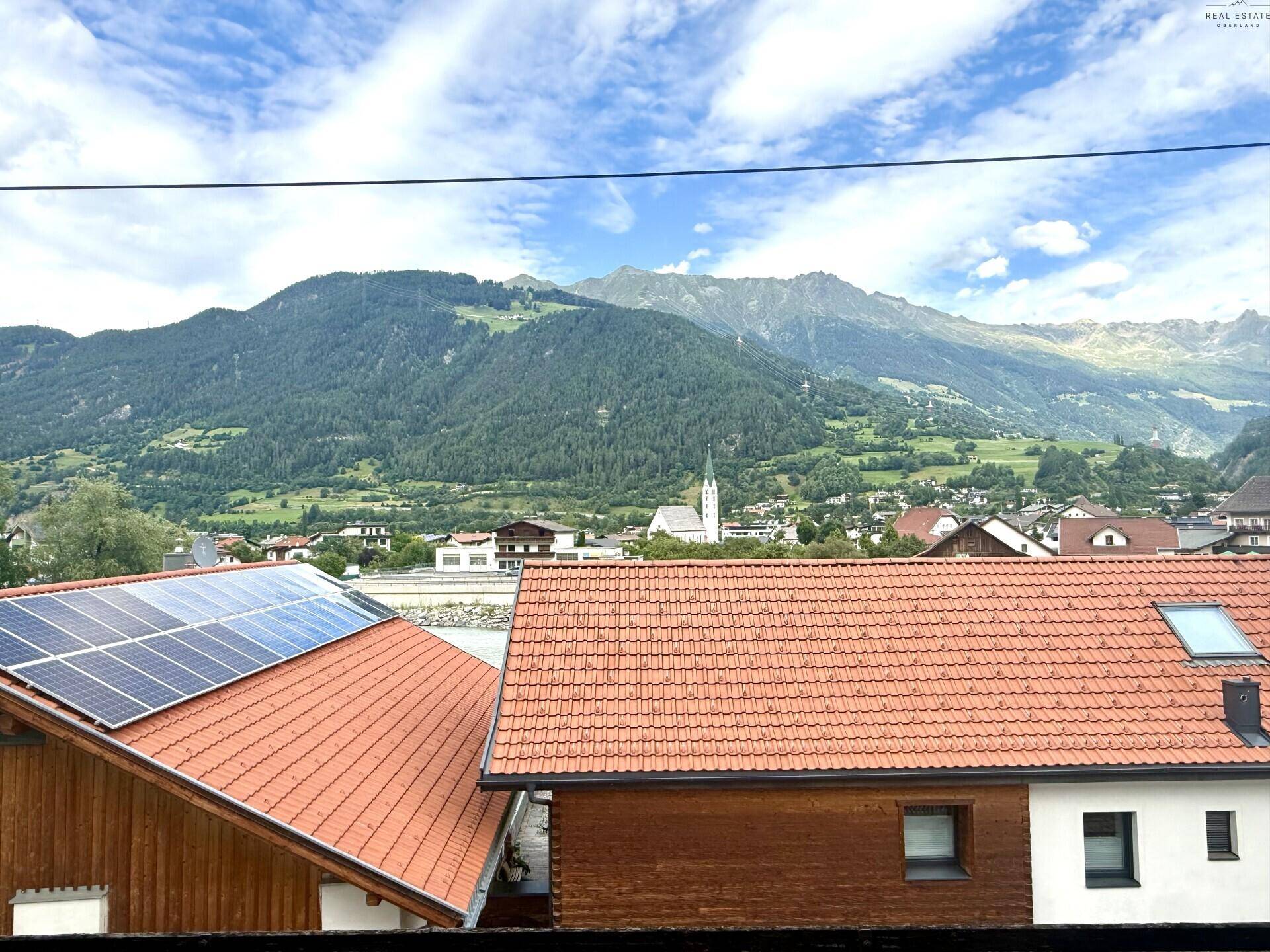 Panoramablick auf die umliegende Berglandschaft mit Dächern und einer Kirche im Tal.