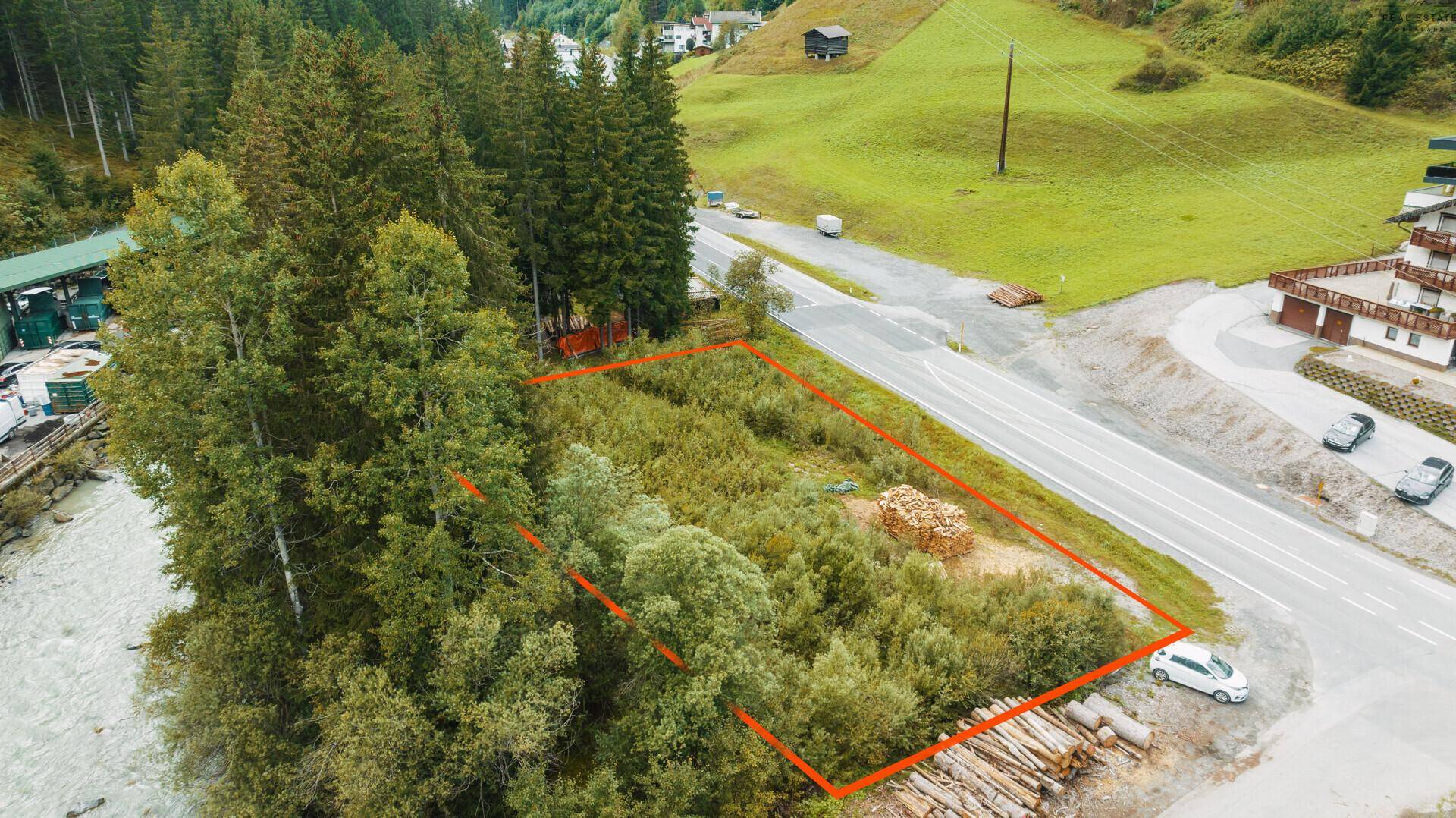 Luftaufnahme eines bewachsenen Grundstücks neben einer Straße und einem Fluss, umgeben von Wald und Gebäuden.